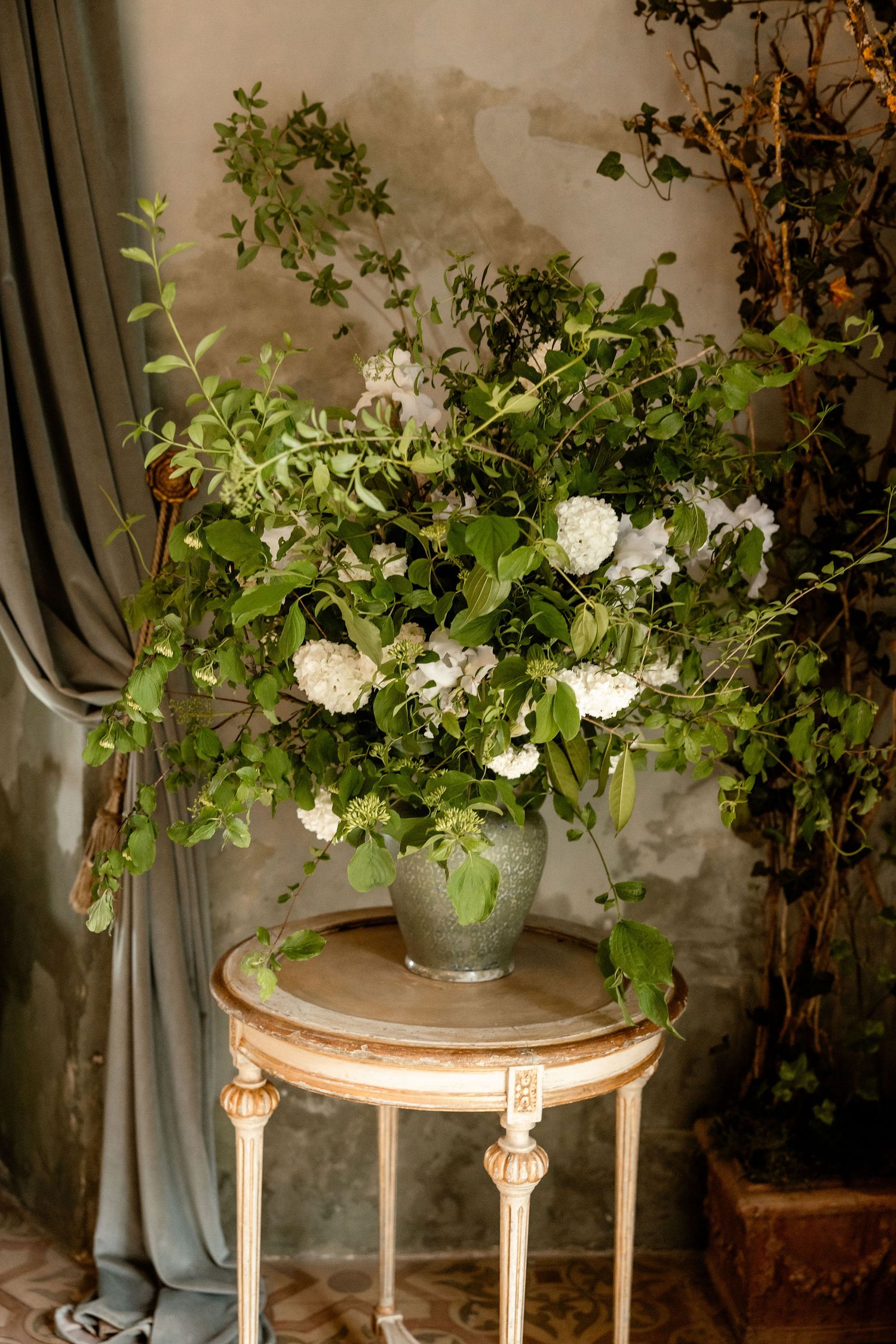 Elegant floral arrangement with white blooms and greenery displayed on vintage ornate side table with blue curtain backdrop.