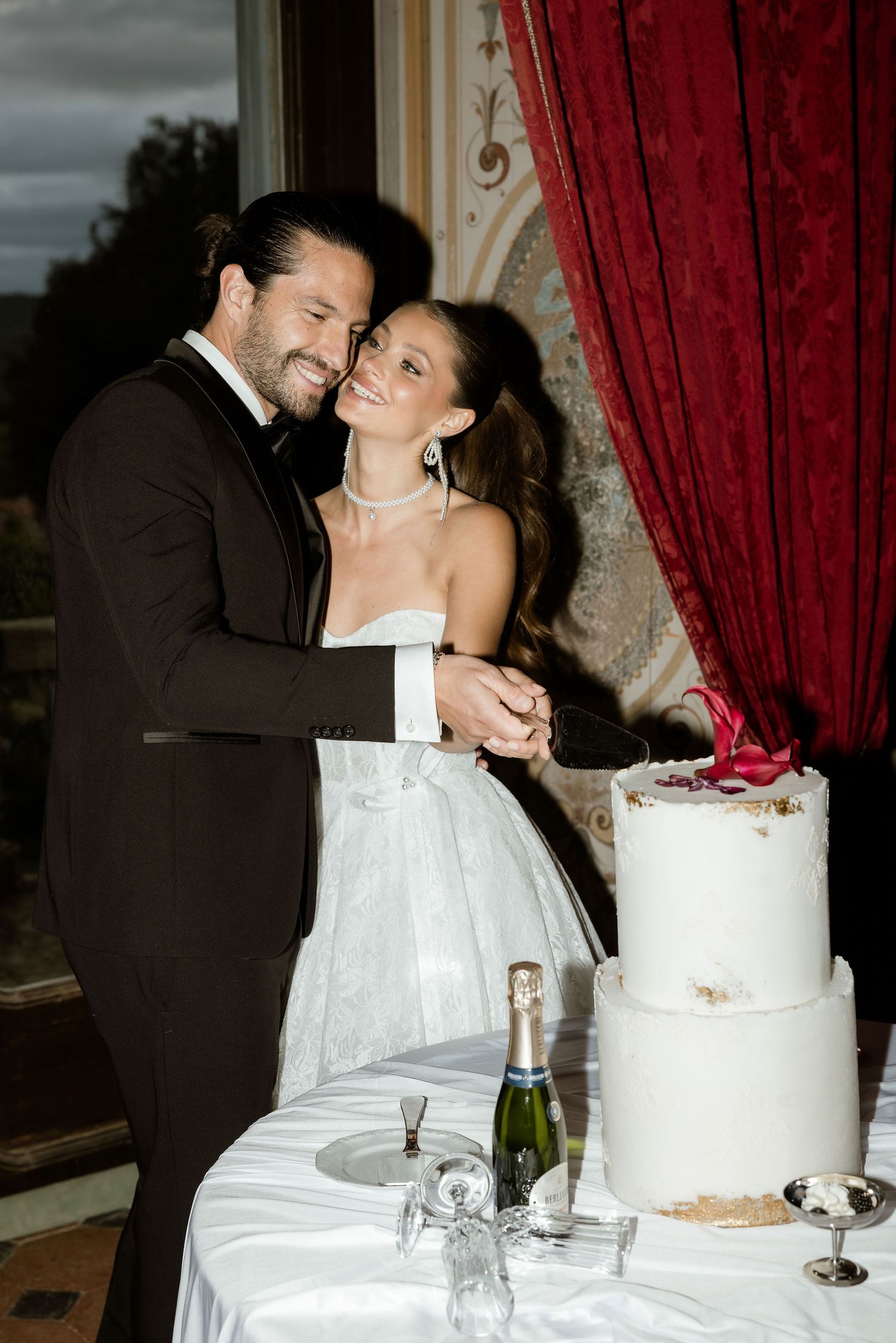 A romantic wedding cake-cutting moment between a couple in formal attire with a champagne bottle nearby.