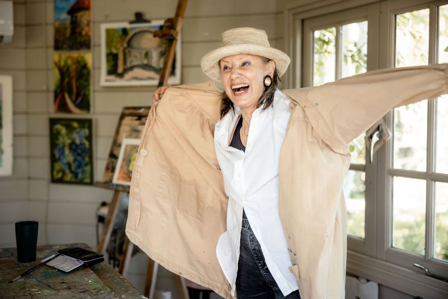 Joyful person in white shirt and tan jacket spreads arms wide while standing in sunlit art studio space.