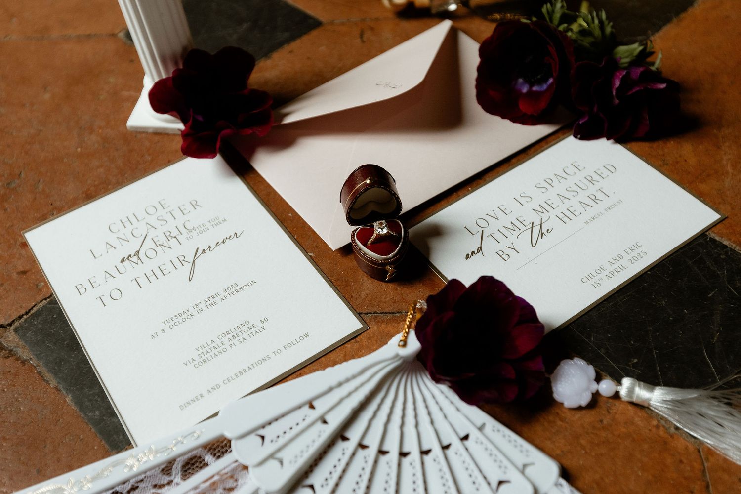 Elegant wedding invitations styled with burgundy flowers and a decorative white fan on a wooden surface.