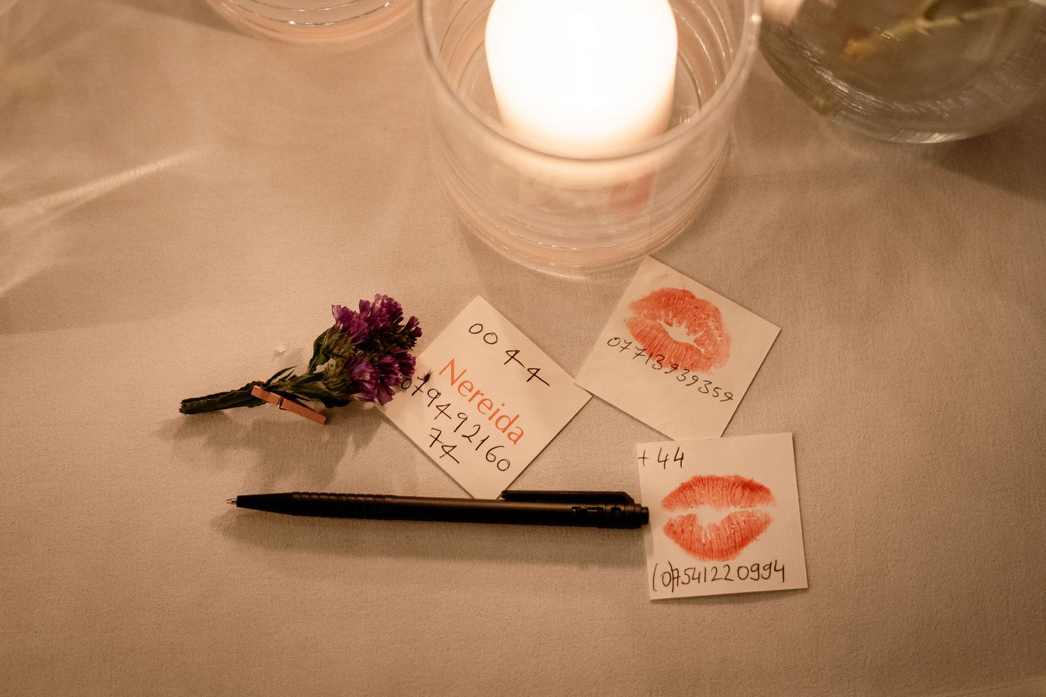 A romantic scene with candlelight, red kiss mark cards, a pen, and a small flower on a white tablecloth.