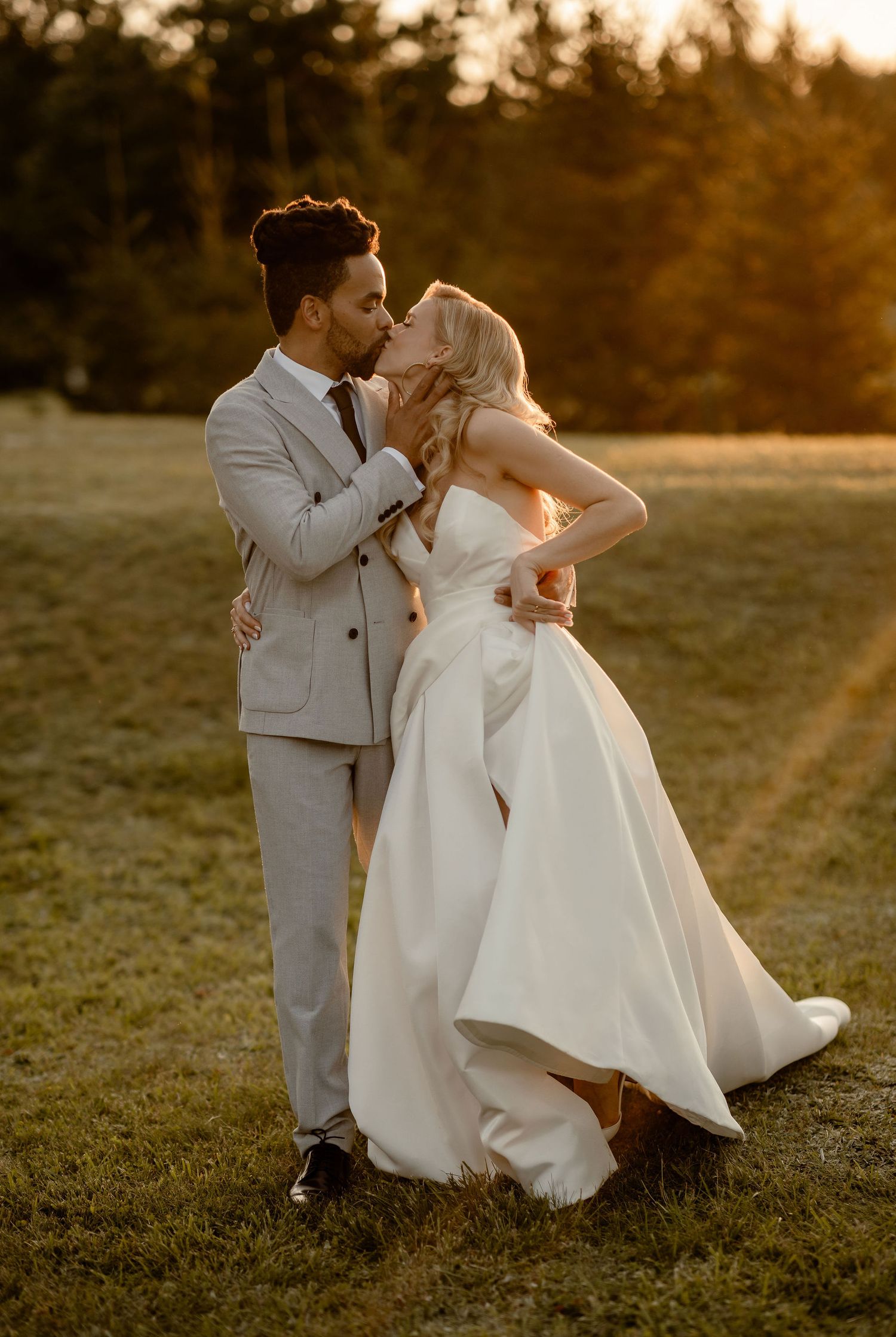 A couple in wedding attire dances together at sunset in a grassy field with trees in the background.