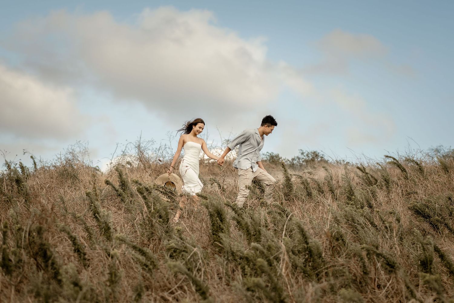 A couple walks through tall grass on a hilltop during golden hour.