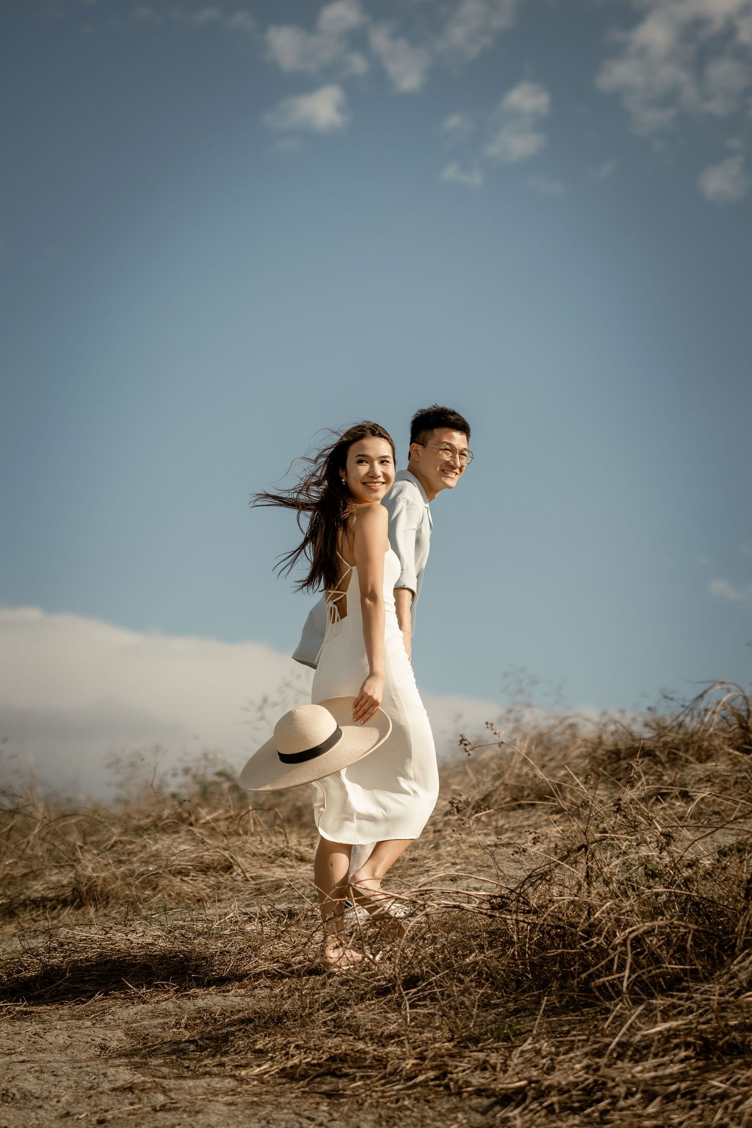 A joyful moment captured on a hilltop with flowing white dress and straw hat against blue sky.