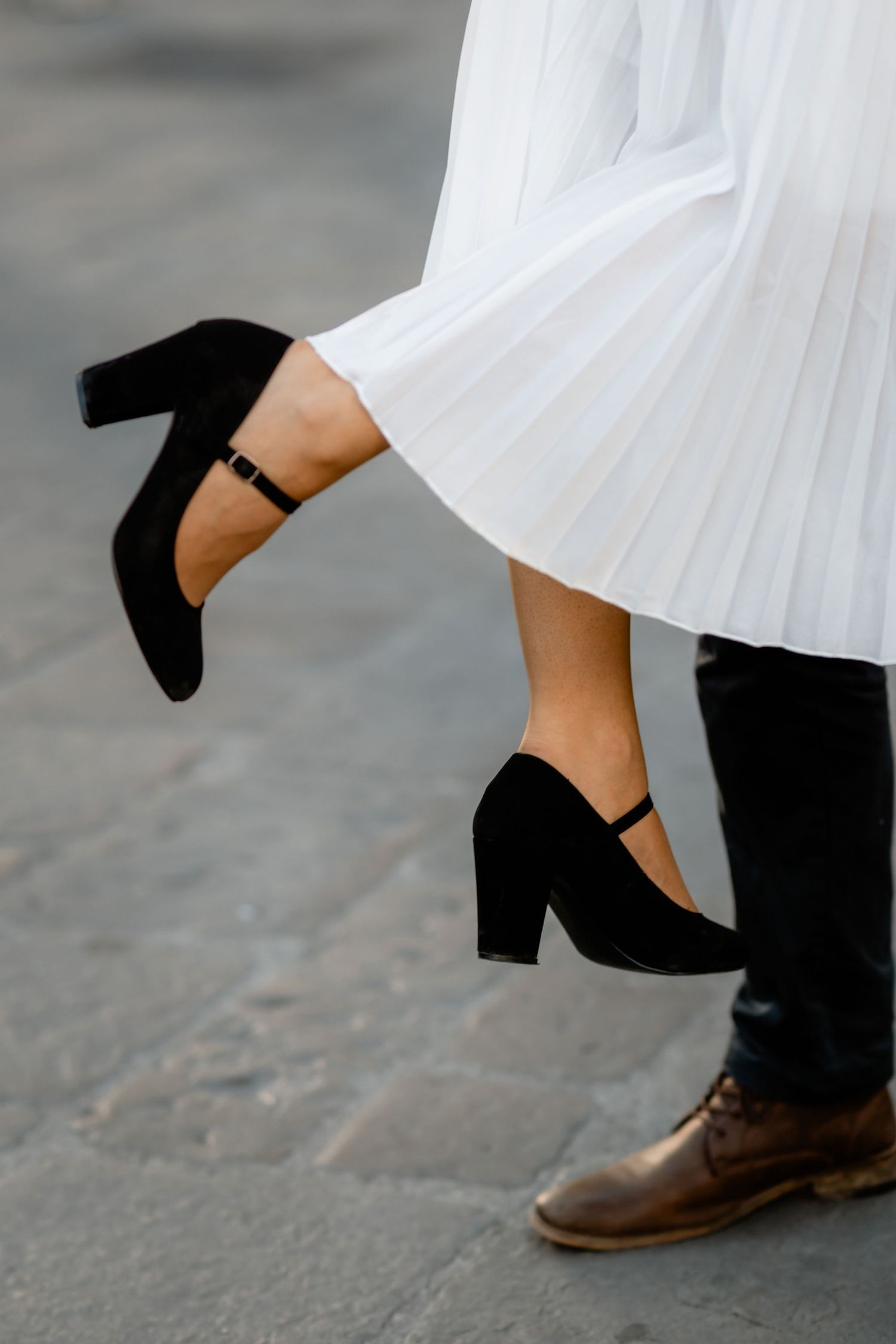 A close-up of contrasting footwear showing a black heeled shoe against a white pleated dress and brown leather shoe.