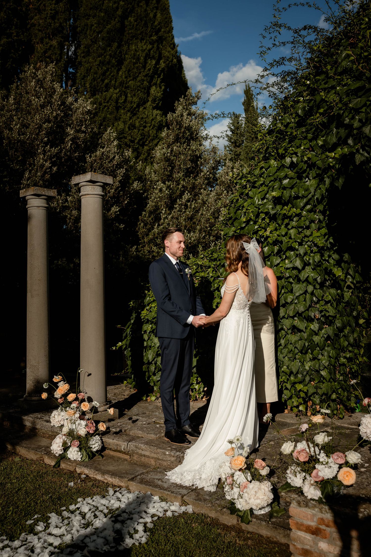 A couple shares an intimate moment under stone pillars surrounded by flowers and greenery.