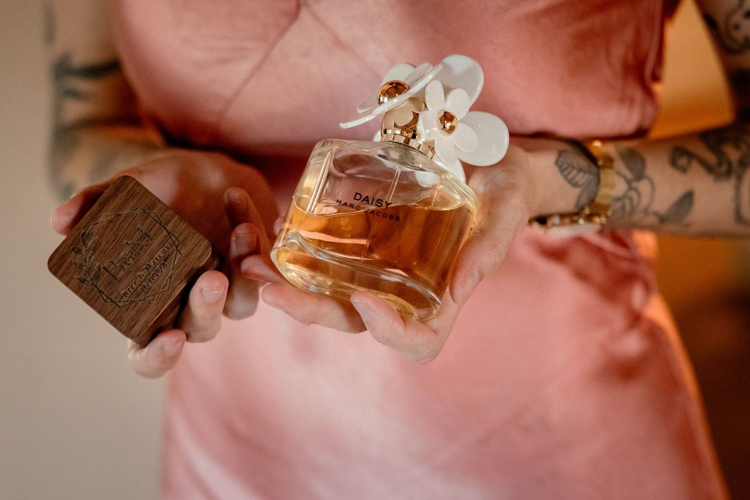 Tattooed hands hold a perfume bottle and wooden box against a pink background.