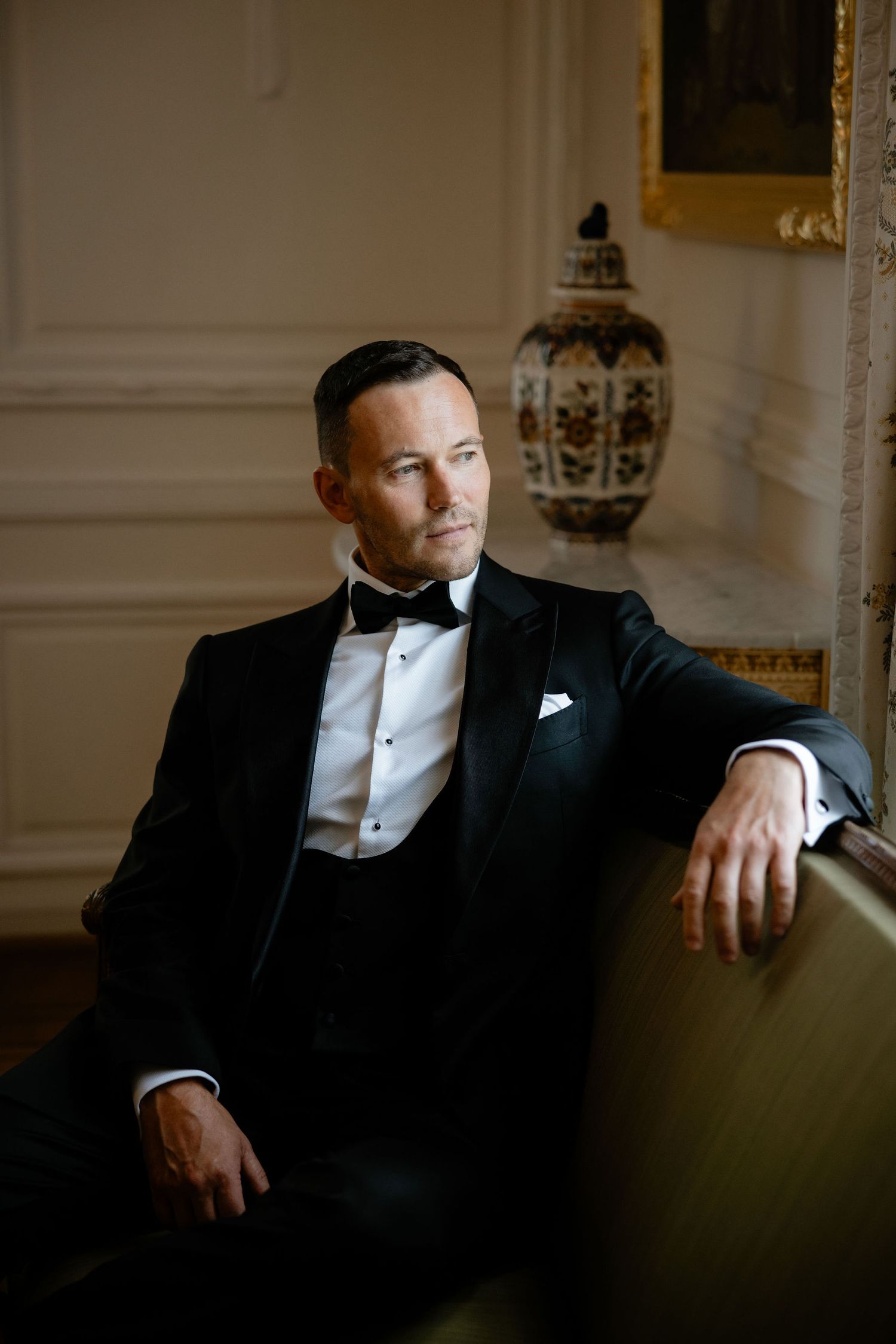 A dapper groom in classic tuxedo poses on vintage furniture in an elegant room.