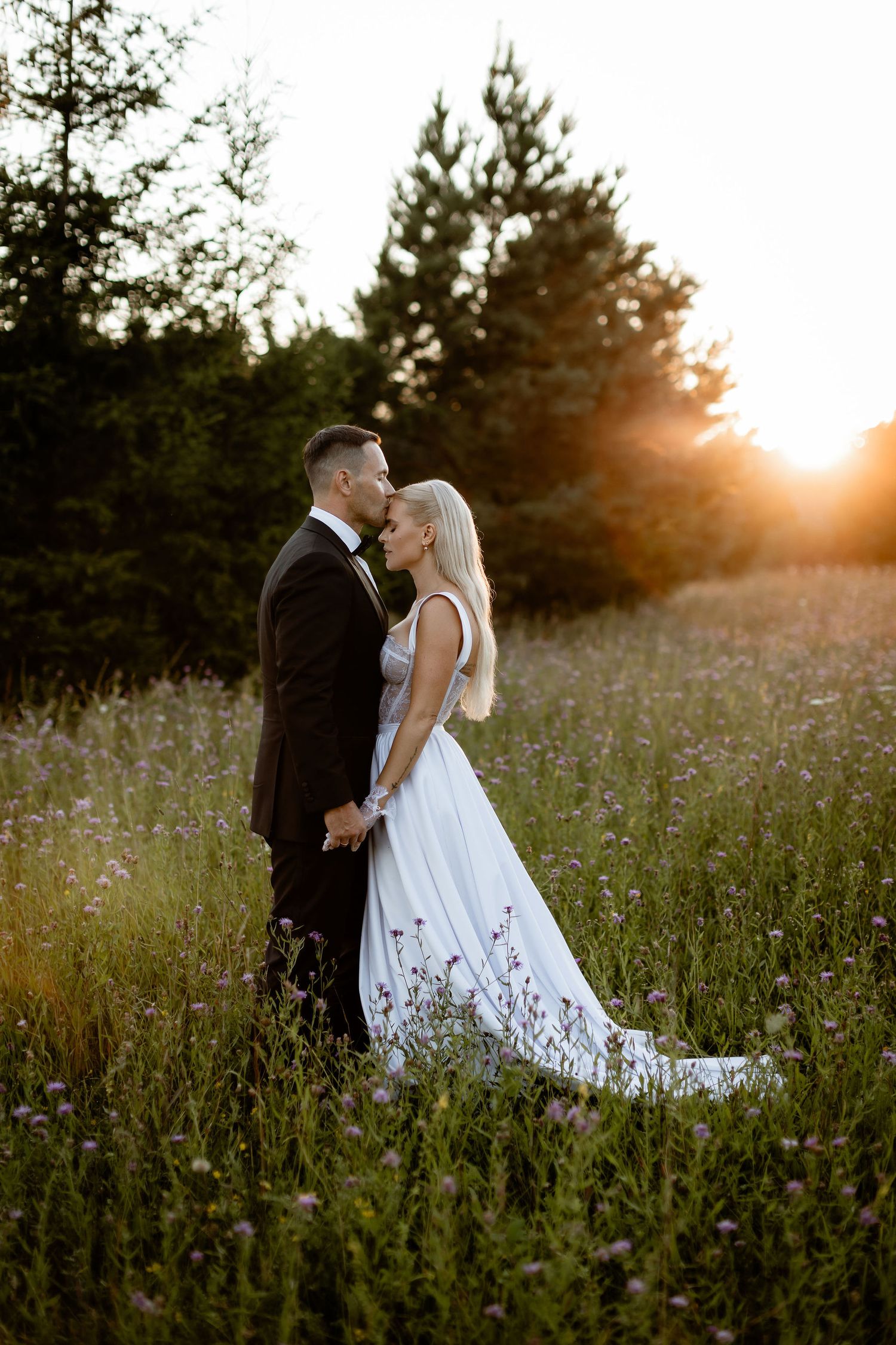 Elegant wedding couple embracing in golden hour light among wildflowers and pine trees.