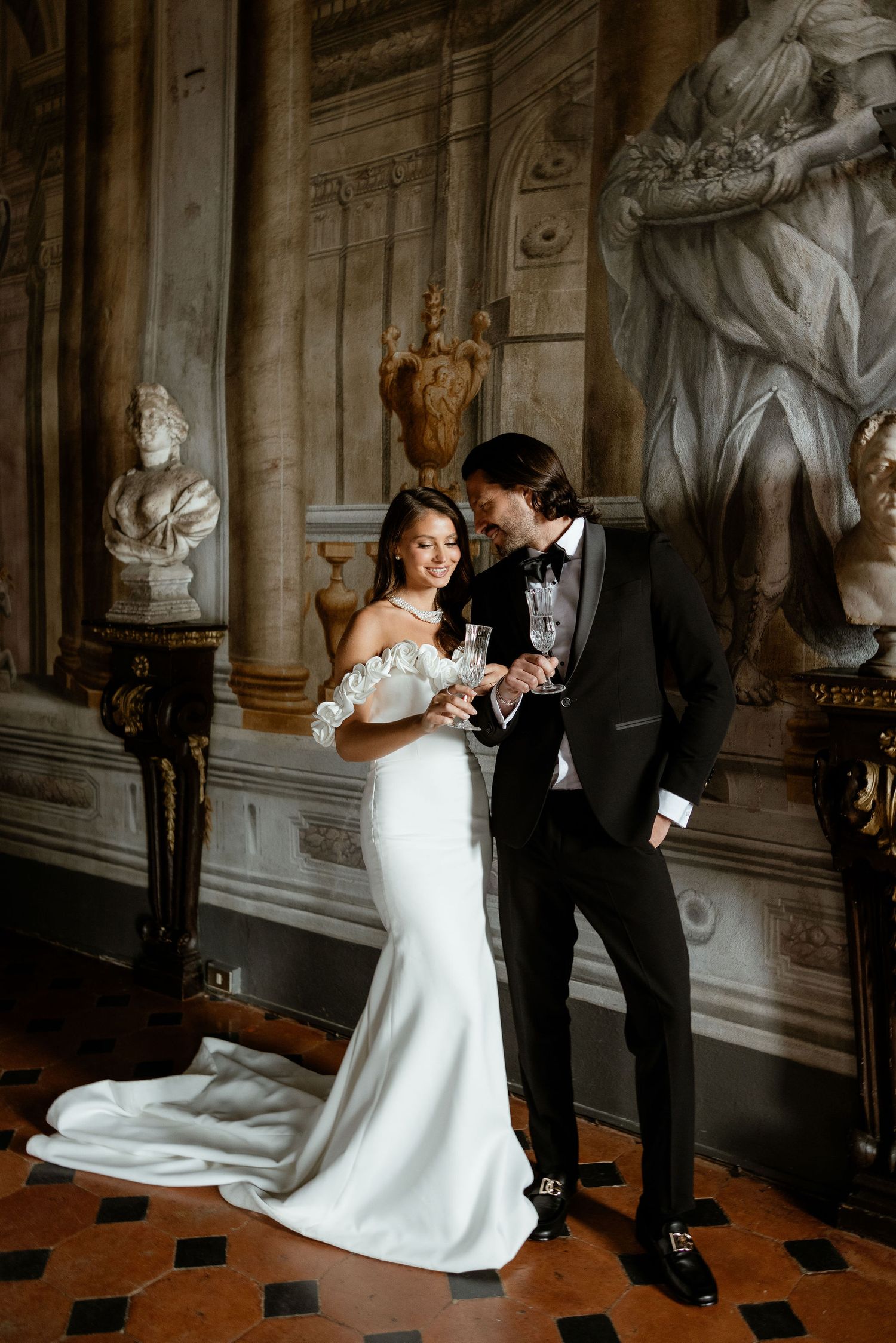 A couple shares a romantic moment in an elegant marble hallway with classical statues and ornate architectural details.