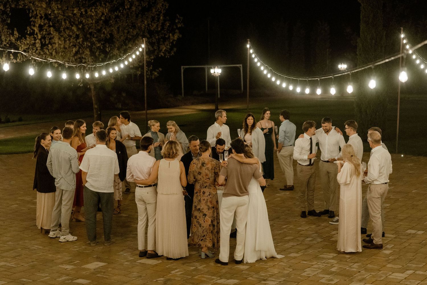 String lights illuminate an outdoor evening wedding reception with guests in white attire mingling on a lawn.