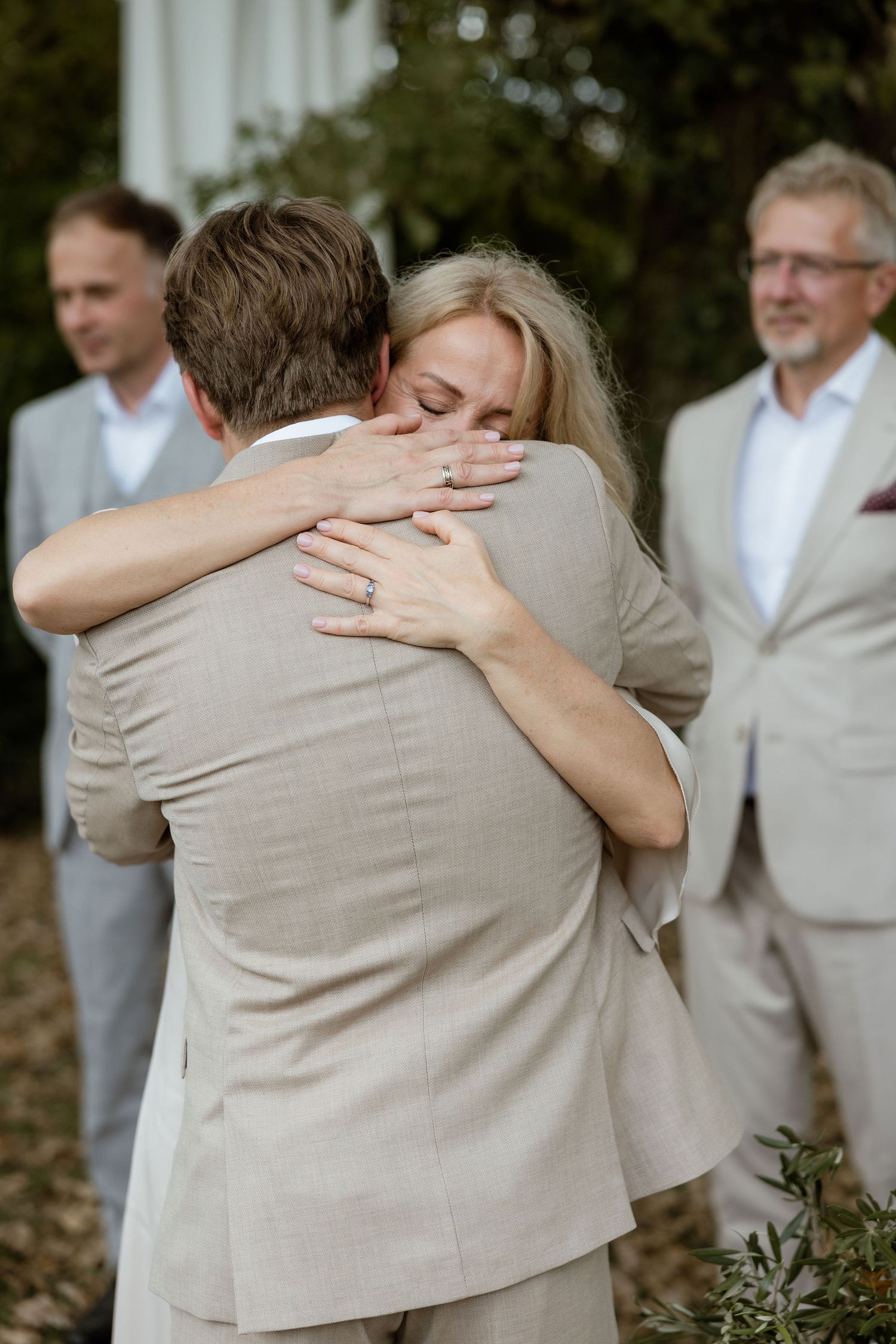 A series of emotional embraces between wedding party members wearing beige and white attire during an outdoor celebration.