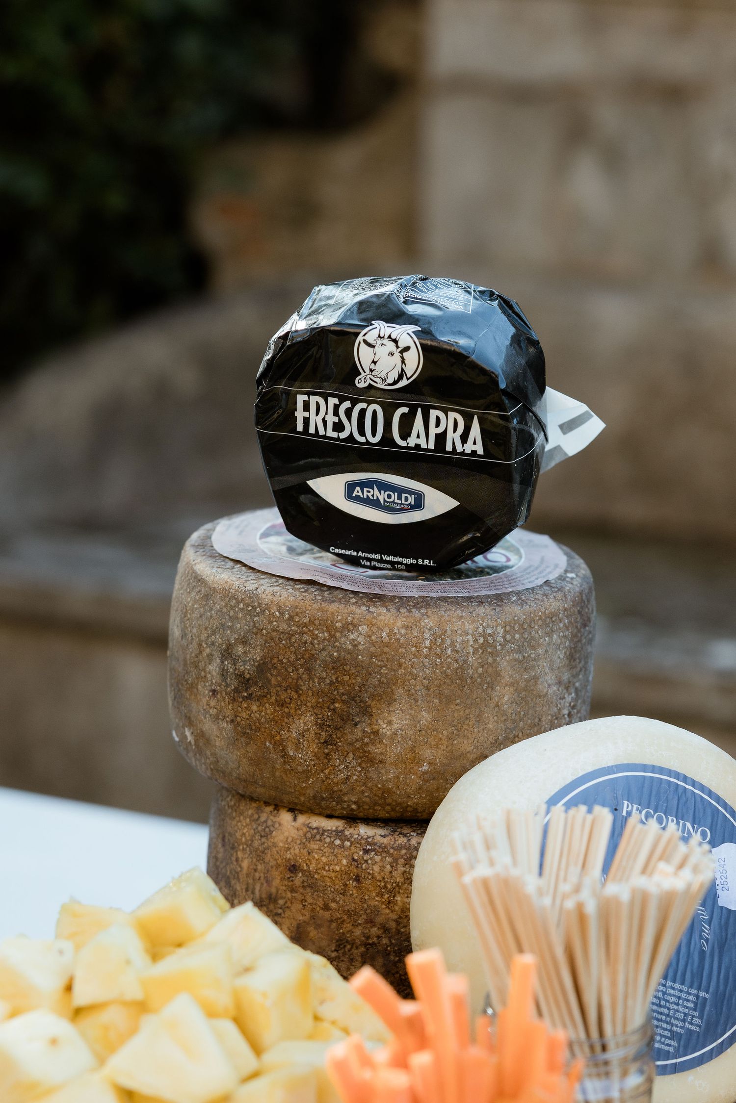 Wheels of aged cheese with black wax tops displayed alongside cubes of fresh cheese on a rustic surface.