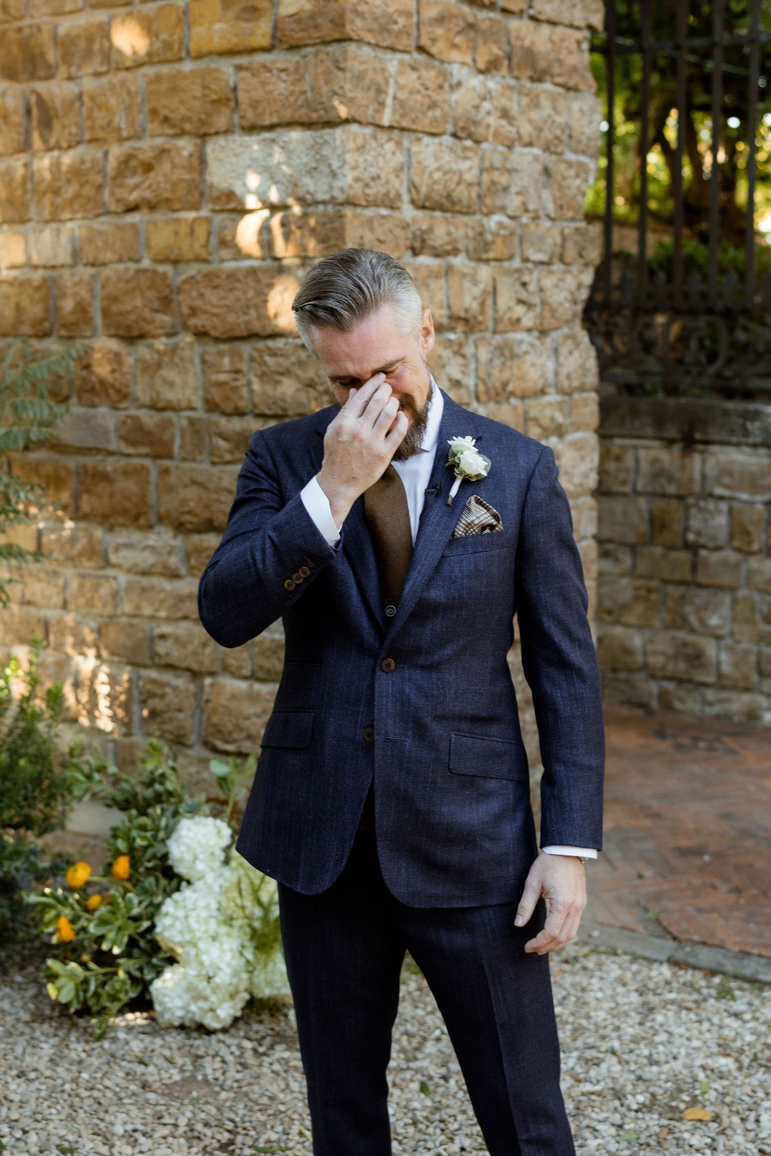 Two people in navy suits share an emotional moment together against a stone wall backdrop at an outdoor wedding venue.