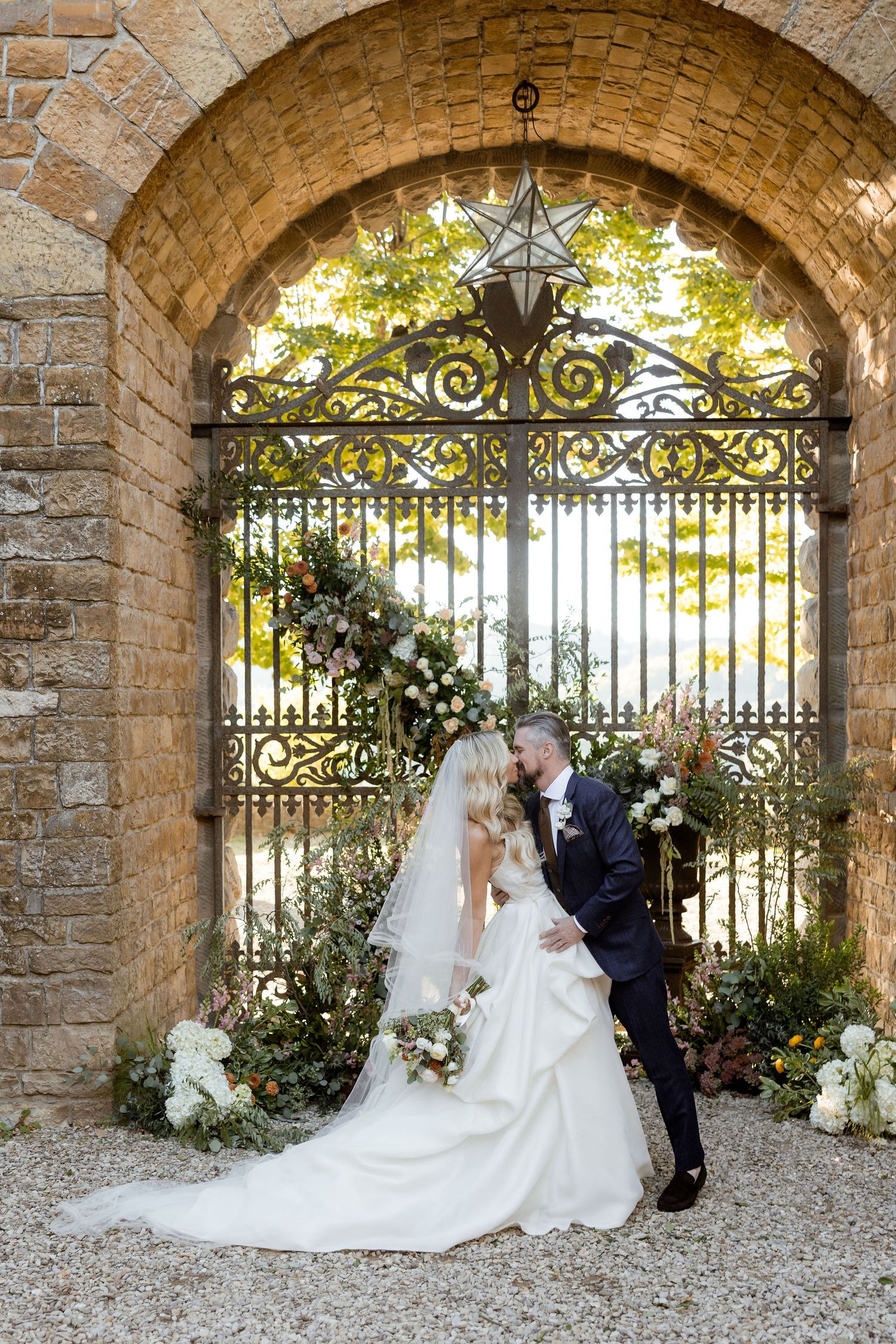 Elegant wedding couple poses under stone archways with ornate wrought iron gates and floral decorations in romantic setting.