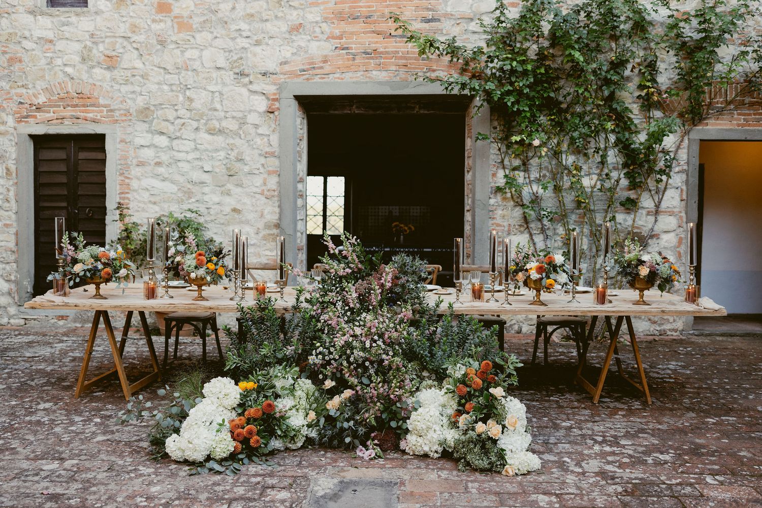 Rustic stone courtyard decorated with elegant floral arrangements and wooden furniture for an outdoor event setup.