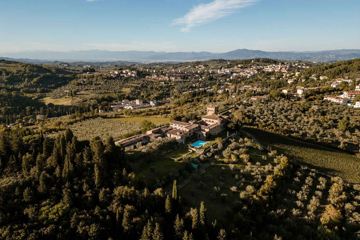 Panoramic aerial view of rolling Tuscan hills dotted with villas and cypress trees stretching to distant mountains.