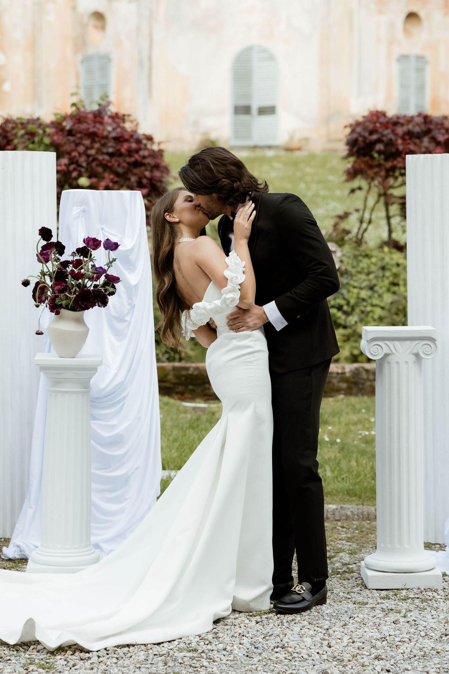 Romantic wedding couple shares a kiss at elegant outdoor ceremony with white columns and purple floral arrangements.
