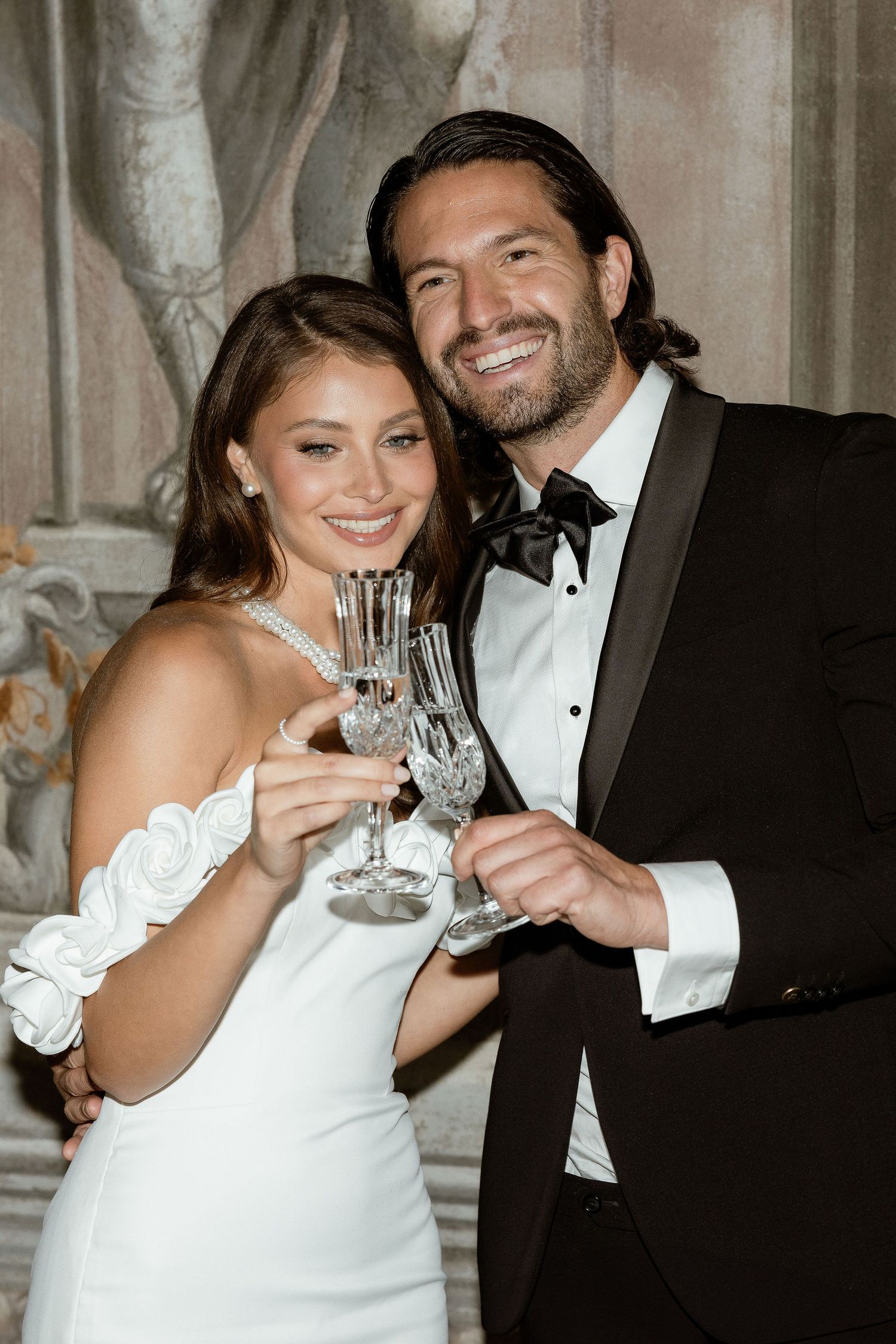 Newlywed couple in wedding attire toasting with crystal champagne glasses at elegant reception venue.