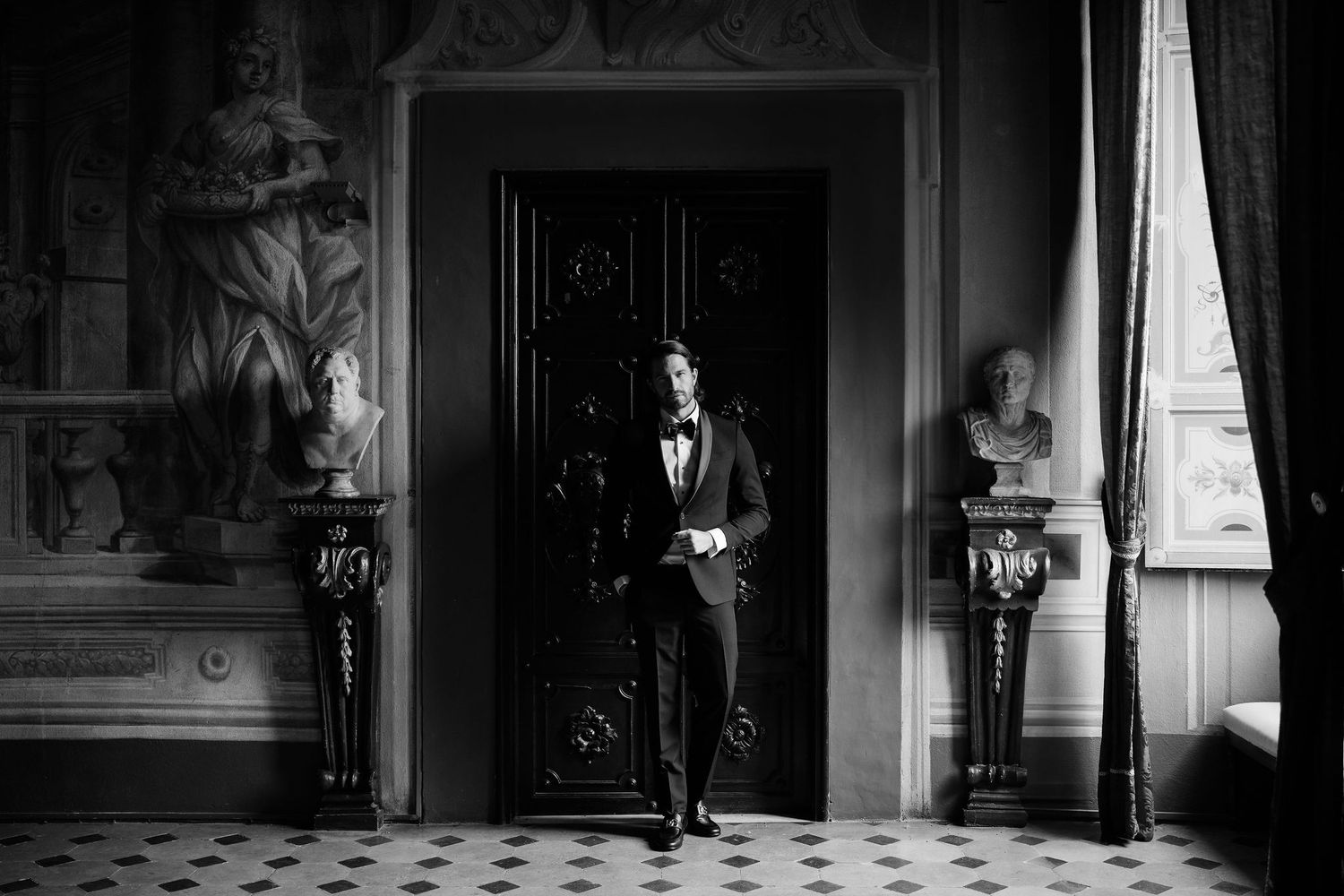 A black and white photograph of an elegantly dressed groom standing between decorative columns in an ornate hallway.