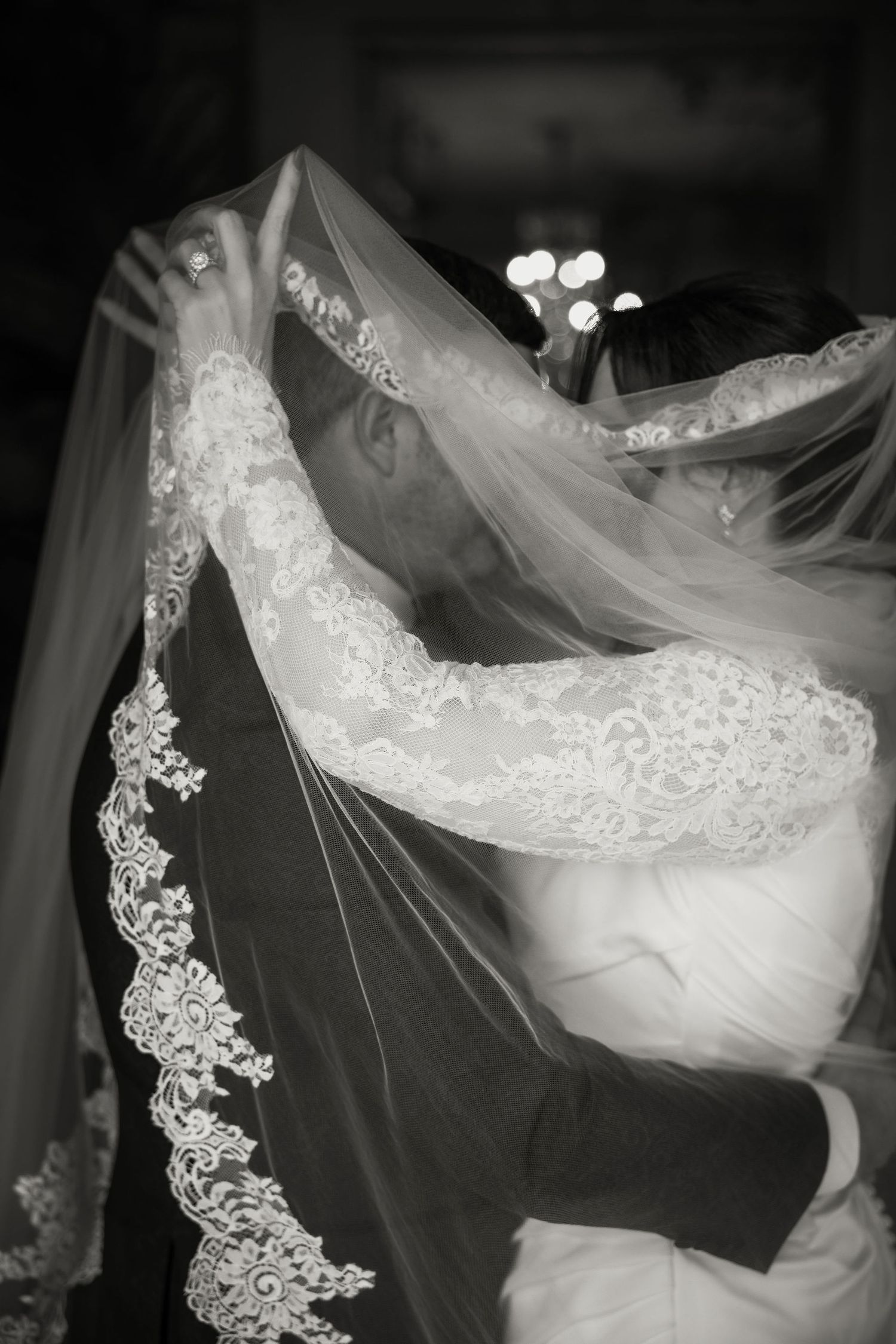 A black and white wedding photograph showing delicate lace veil details draped elegantly over an embracing couple.