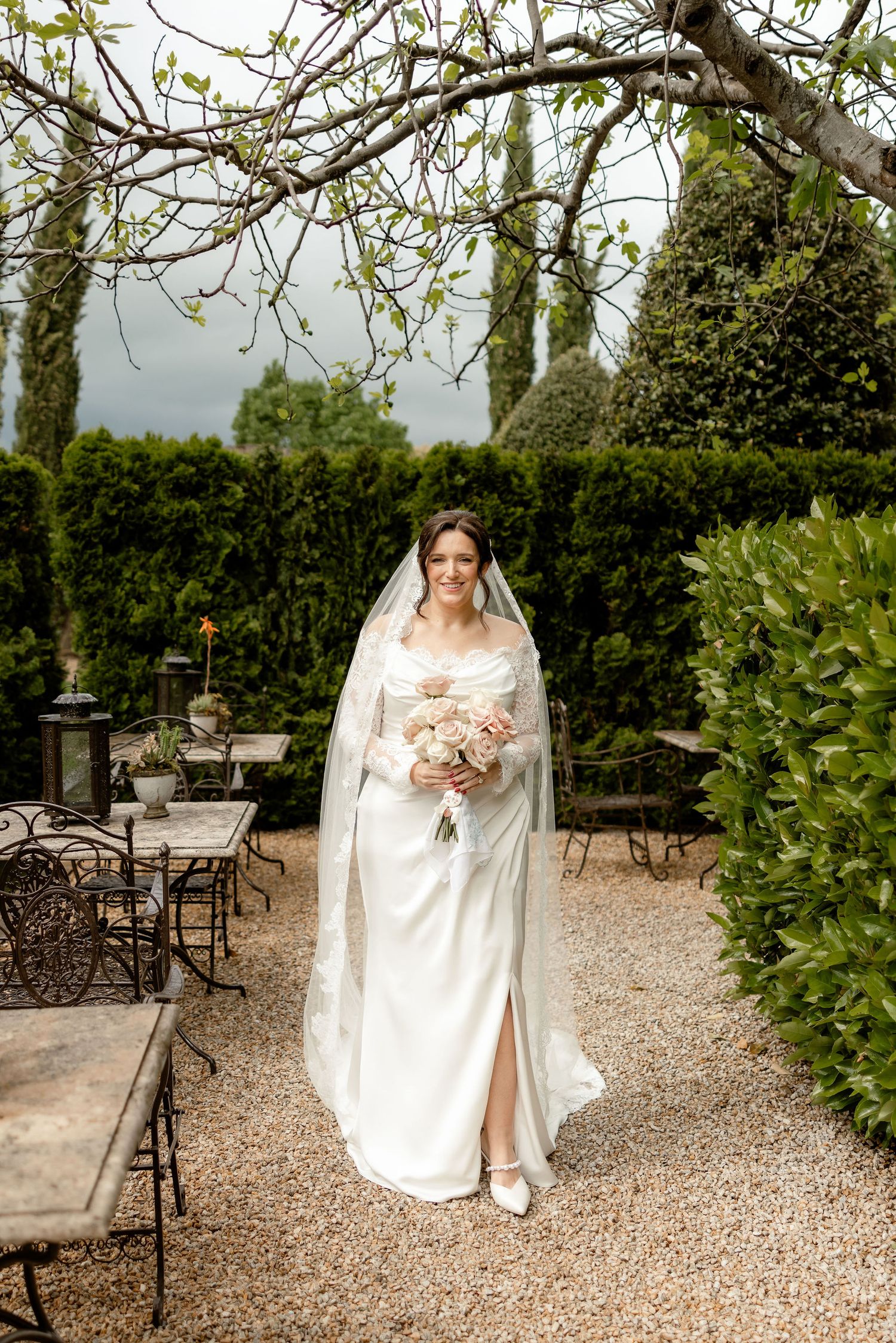 A bride in an elegant white gown and veil stands in a lush garden courtyard with gravel paths and iron furniture.