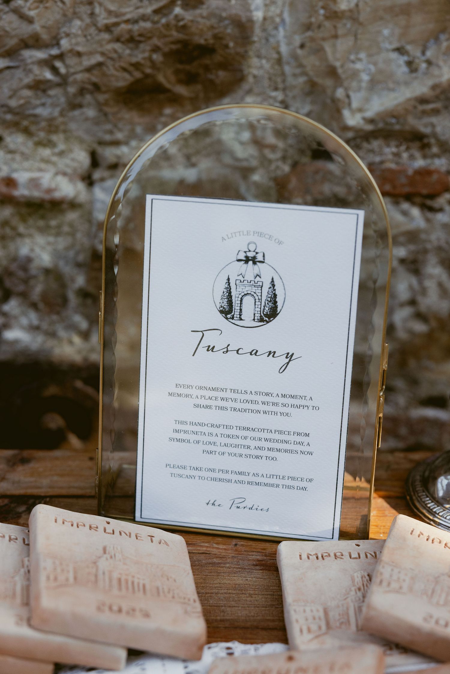 Elegant wedding menu card with Tuscany theme displayed in curved glass frame against rustic stone wall backdrop.