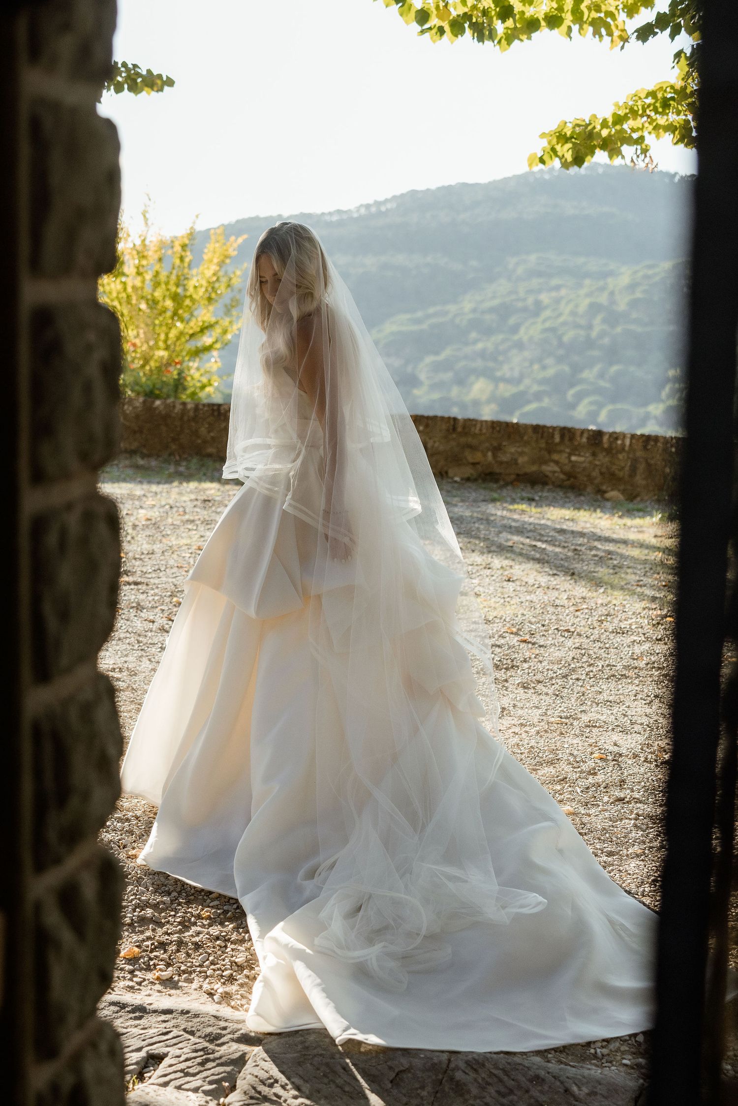 A bride in a flowing white wedding gown and veil stands on a stone terrace overlooking scenic mountain views.
