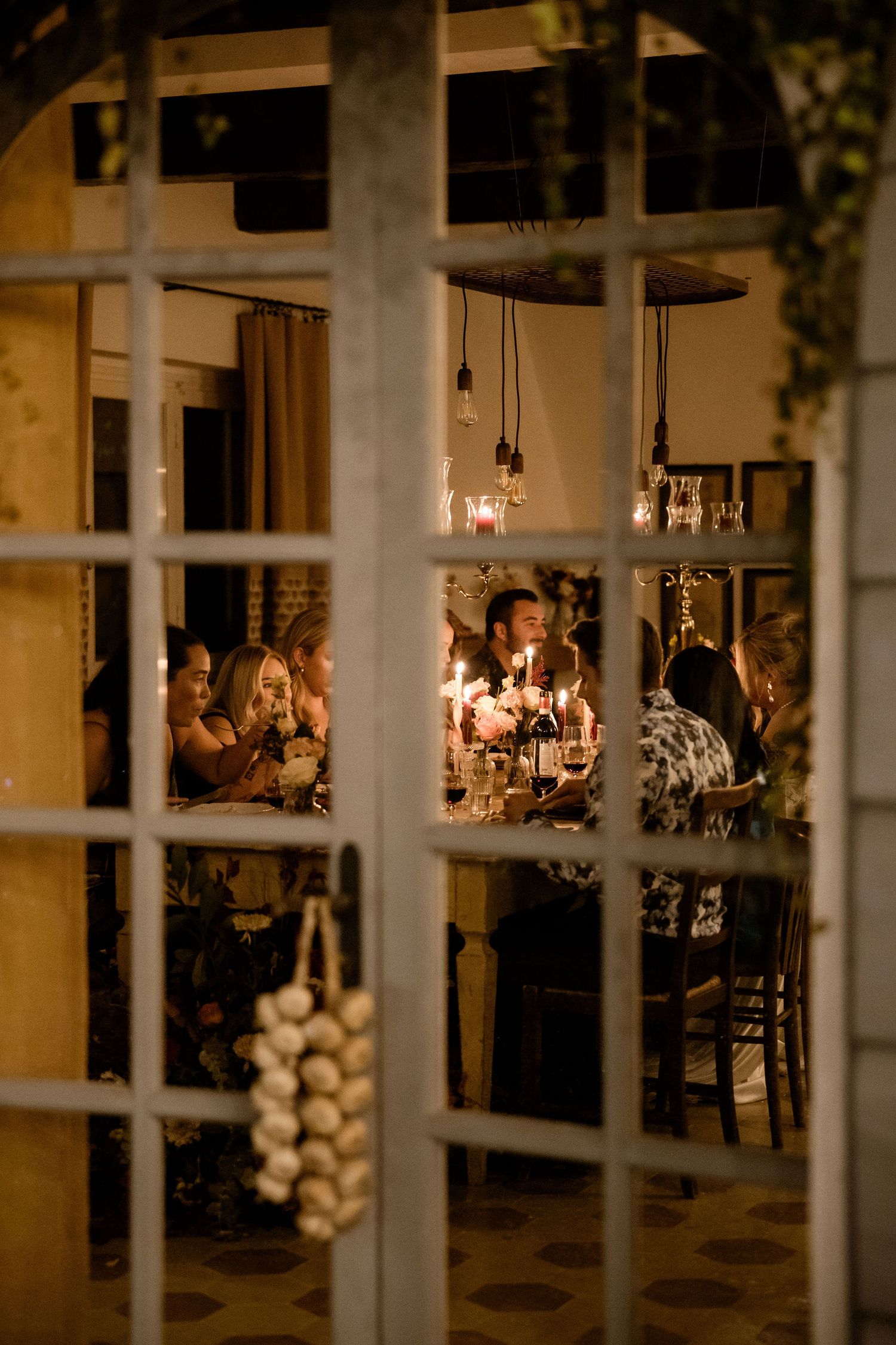 Candlelit dinner party viewed through window panes with glowing ambiance and hanging pendant lights.