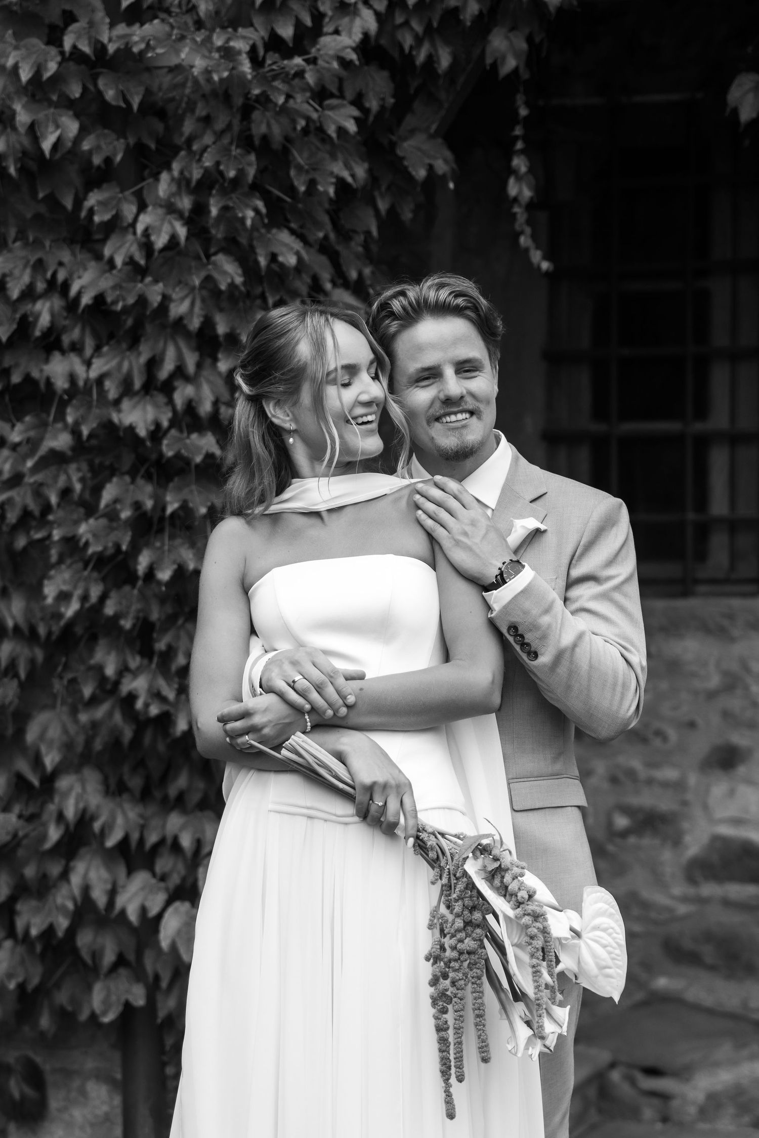 A black and white wedding photo showing a couple embracing and laughing together against an ivy-covered wall.