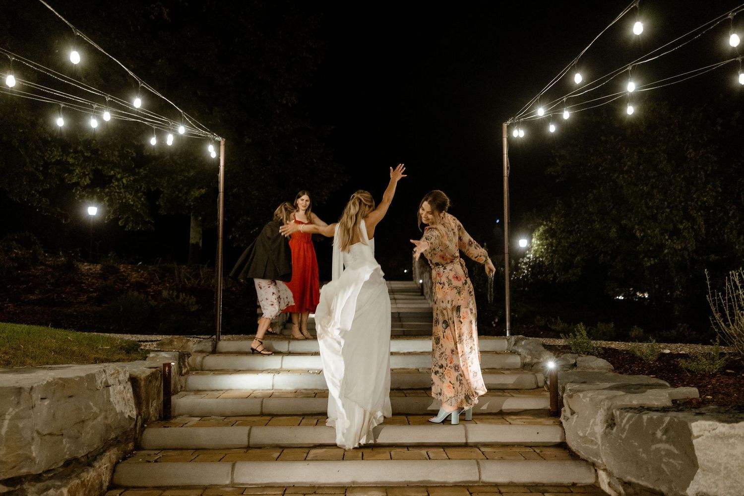 People celebrating at night on stone steps under romantic string lights at an outdoor venue.