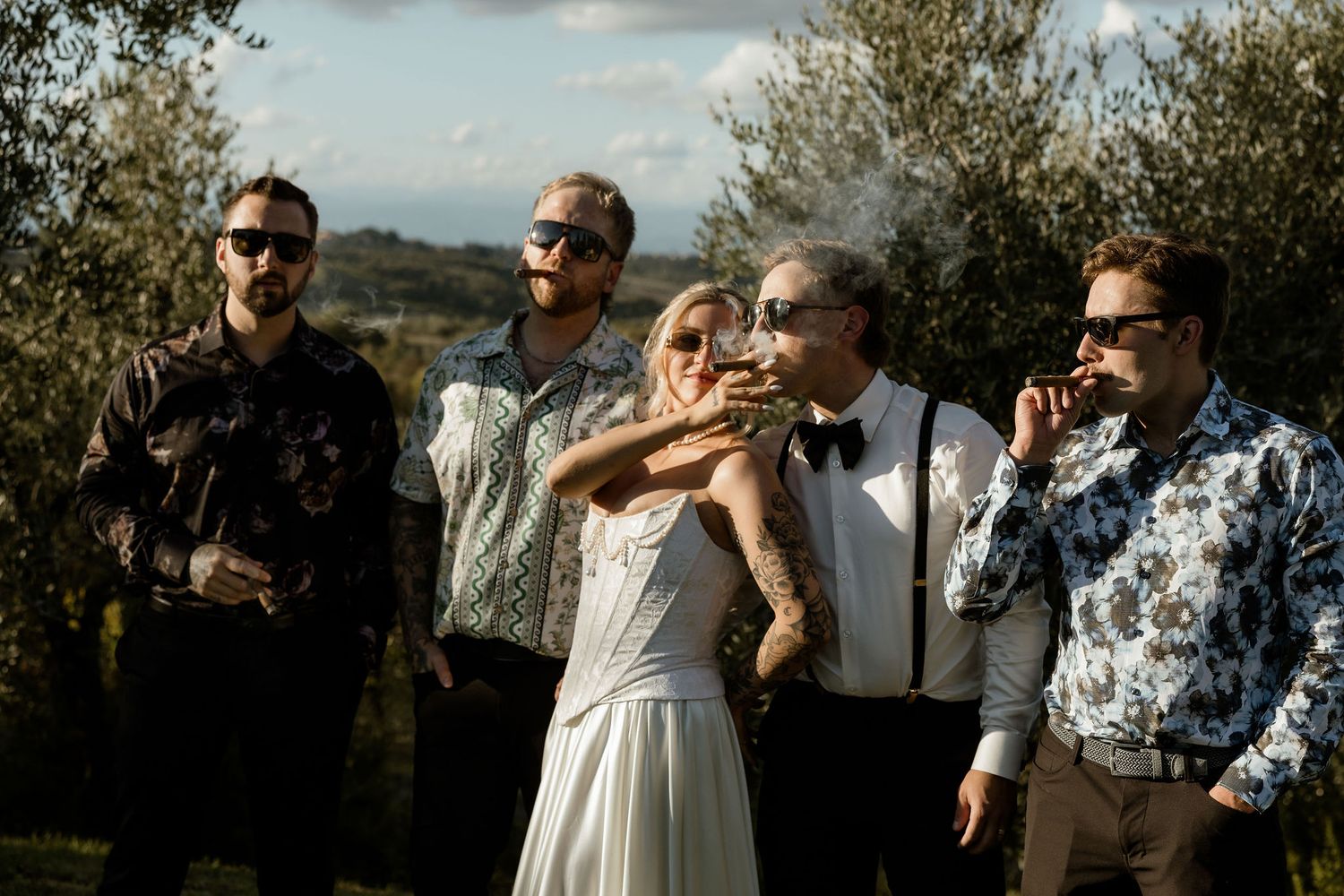 Outdoor wedding celebration with guests dancing and mingling under a sunny sky with trees in the background.