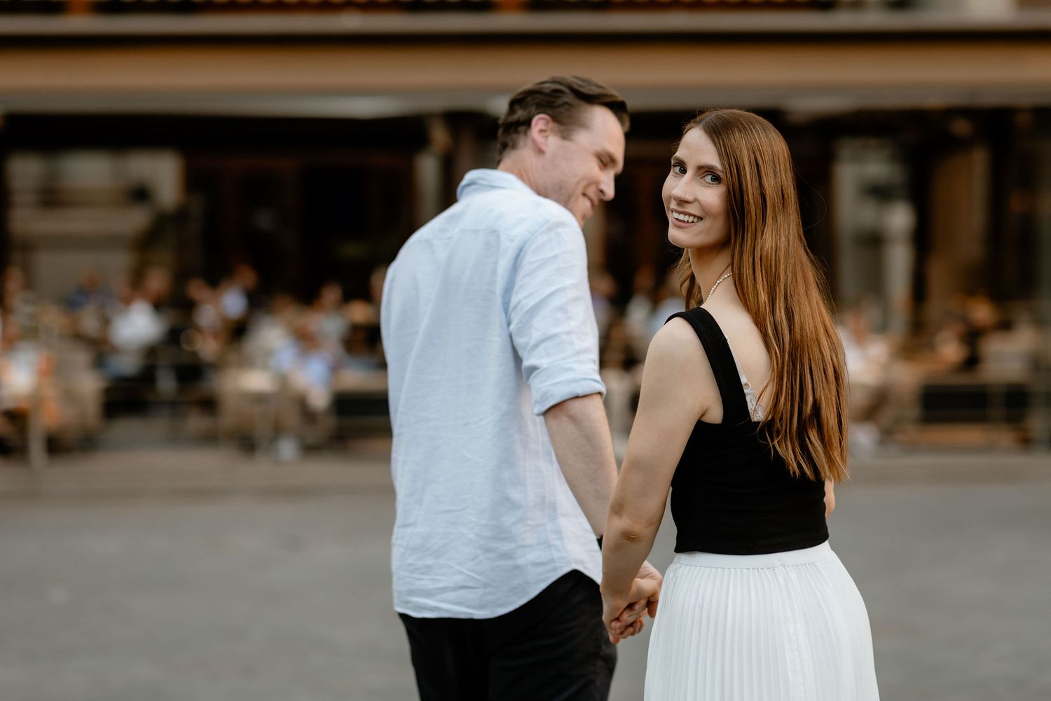 A happy couple holds hands and smiles at each other while standing in front of an outdoor cafe.