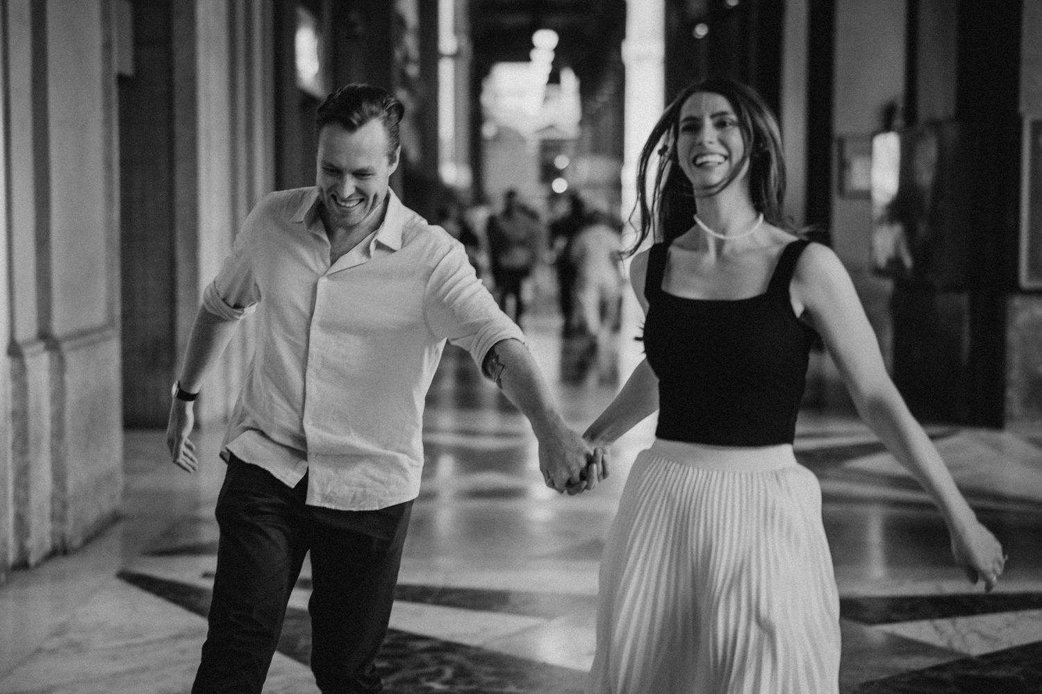 A couple holds hands while walking playfully through an elegant corridor in black and white.