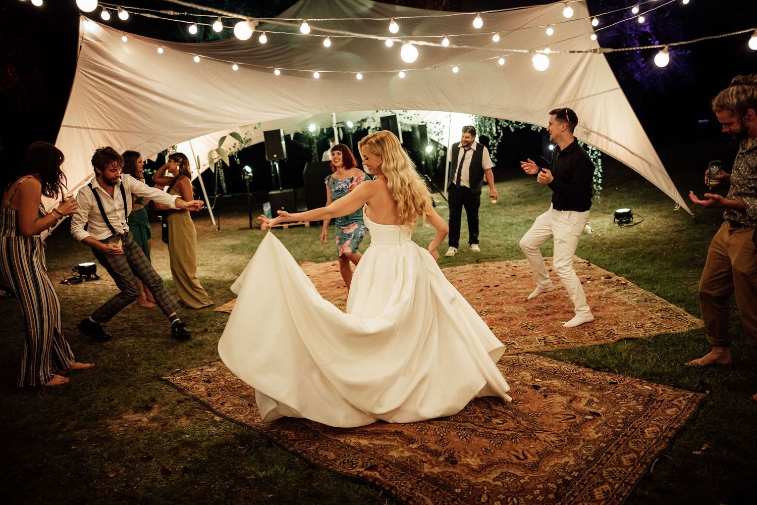 A joyful dance scene unfolds under string lights at an outdoor wedding reception on a rustic dance floor.