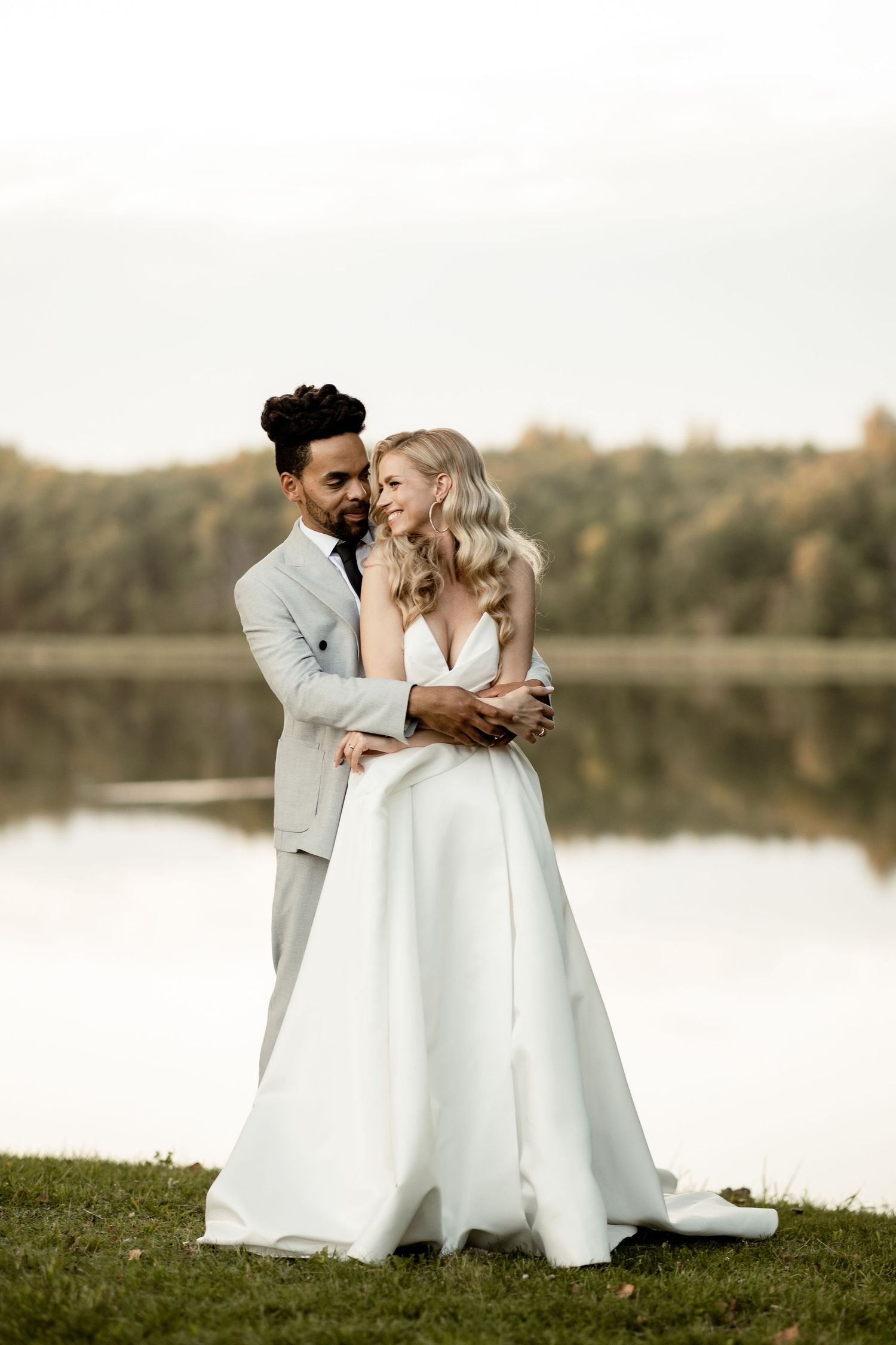 A romantic wedding couple embraces by a serene lake at sunset with autumn trees reflecting in the calm water.