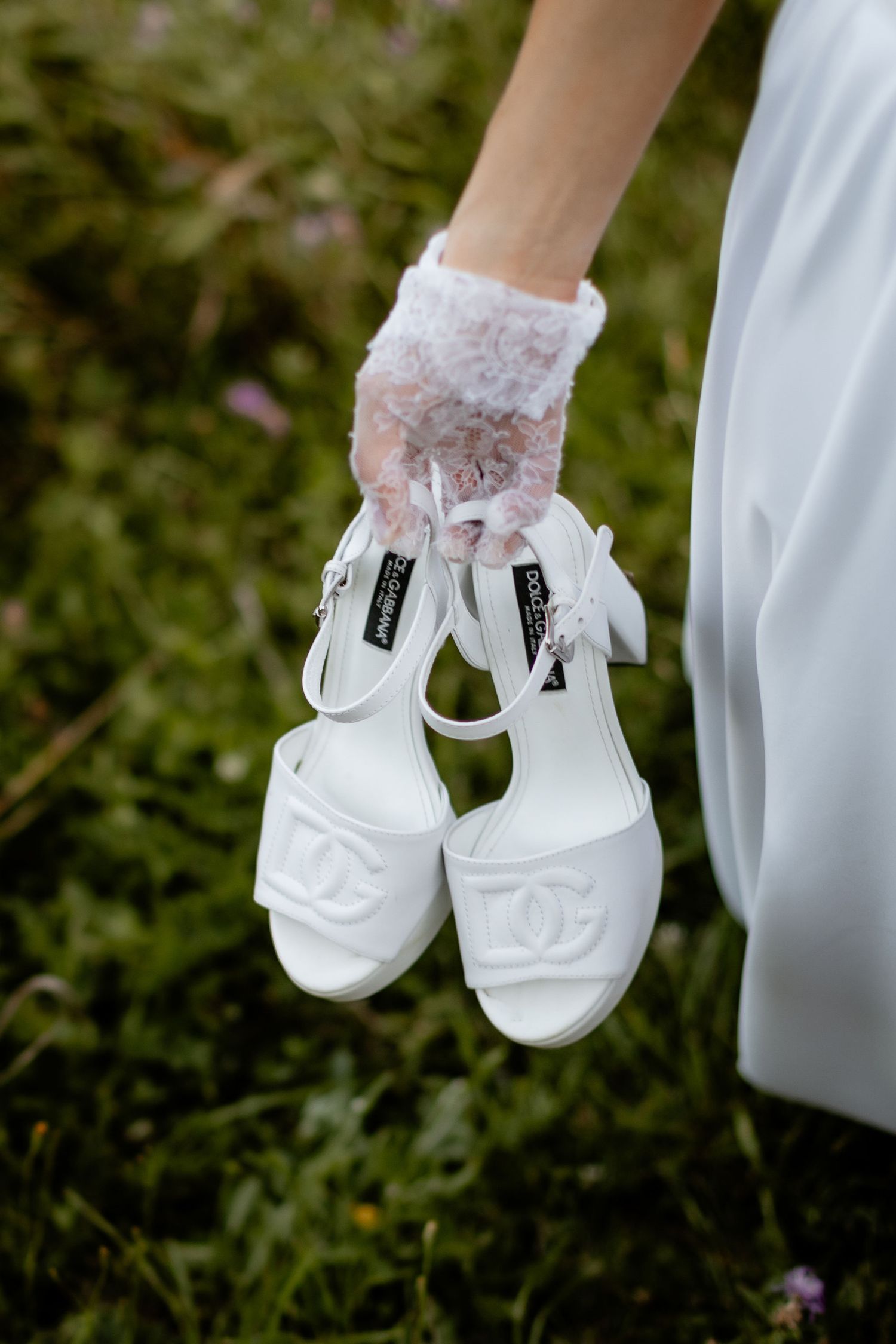 White lace bridal socks and elegant wedding shoes held against greenery background.