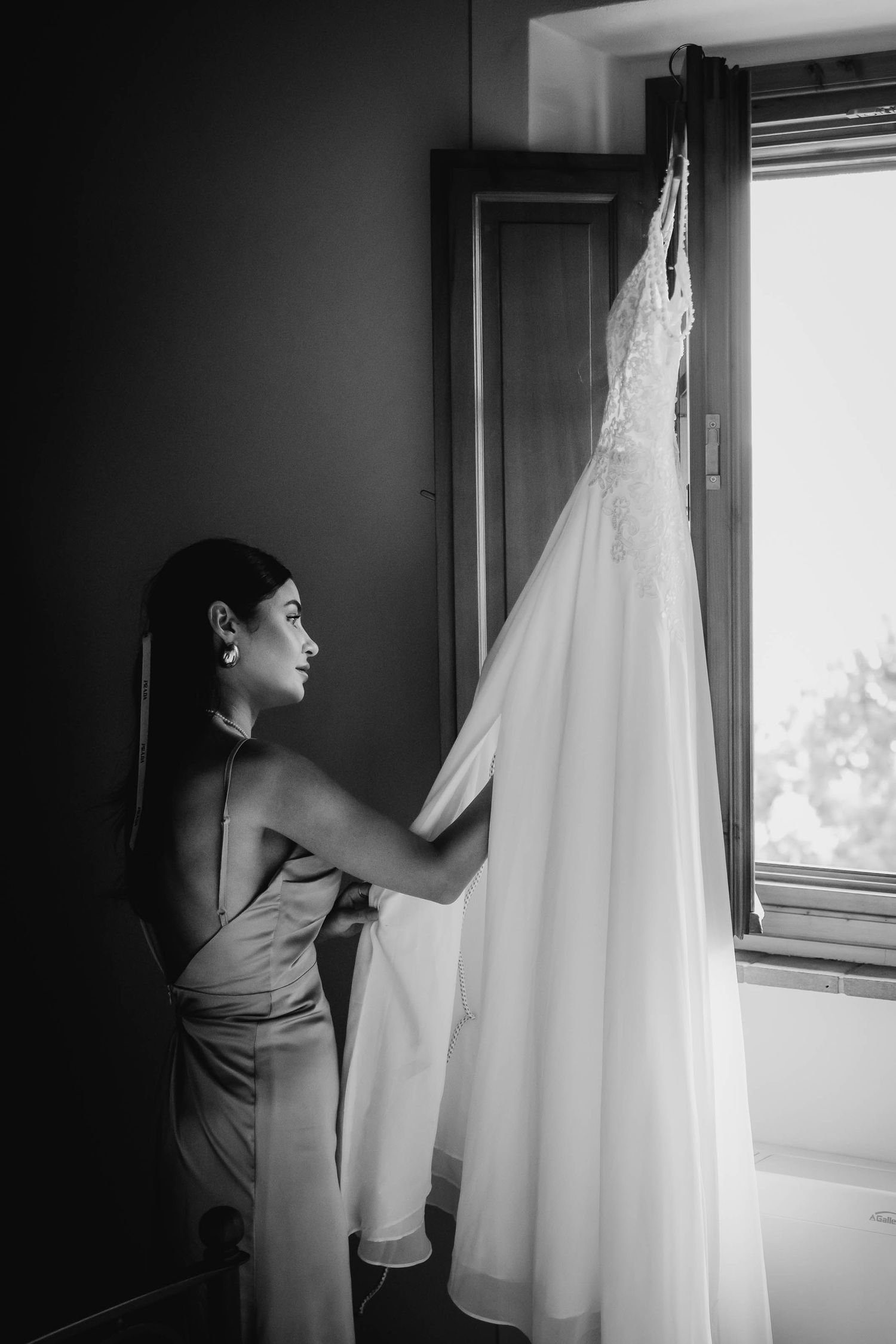 Black and white photo of a wedding dress hanging by a window as someone admires it.