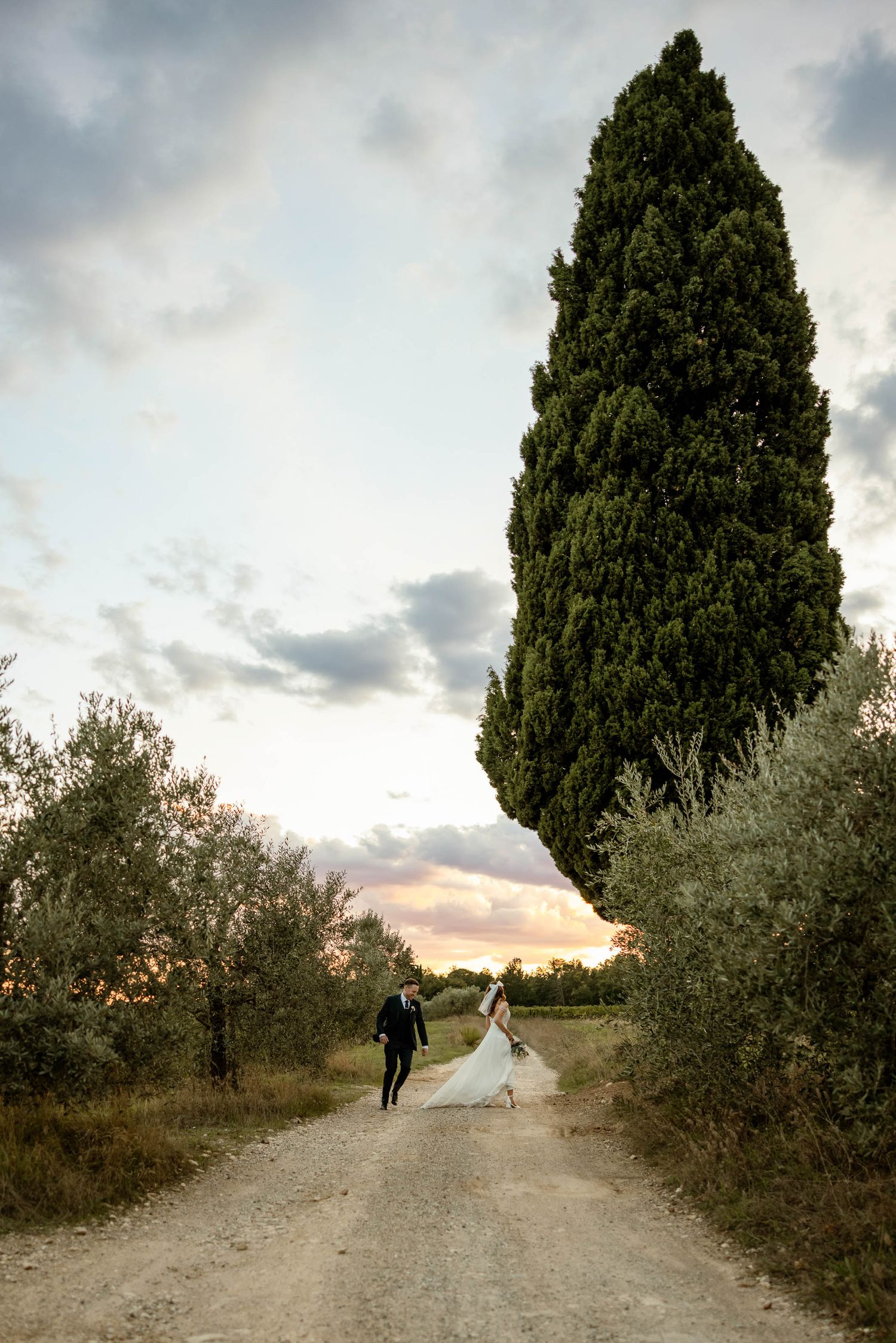 A romantic couple walks down a cypress-lined dirt path in Tuscany at sunset.