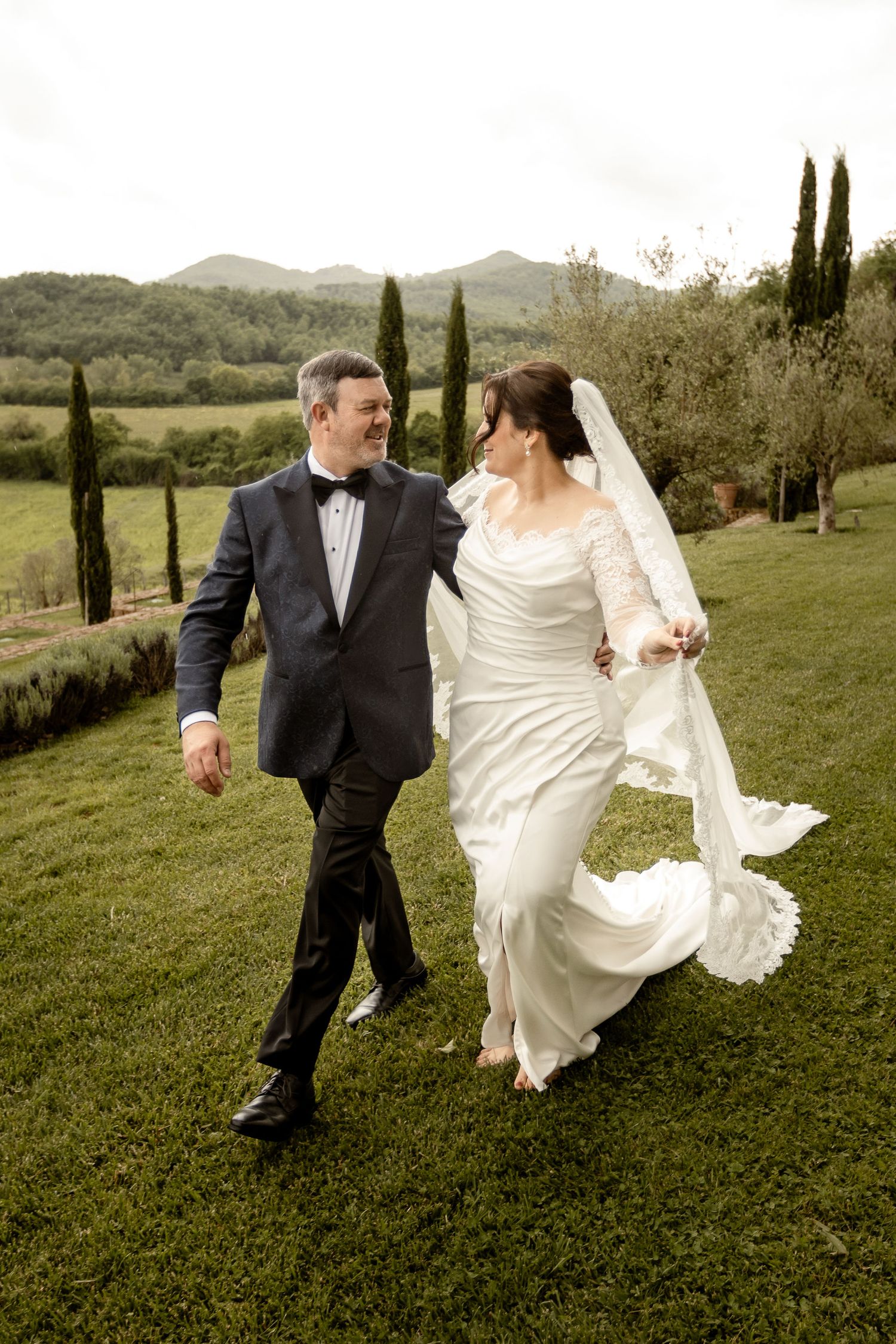 A wedding couple walking on lush green grass with cypress trees and mountains in the Italian countryside background.
