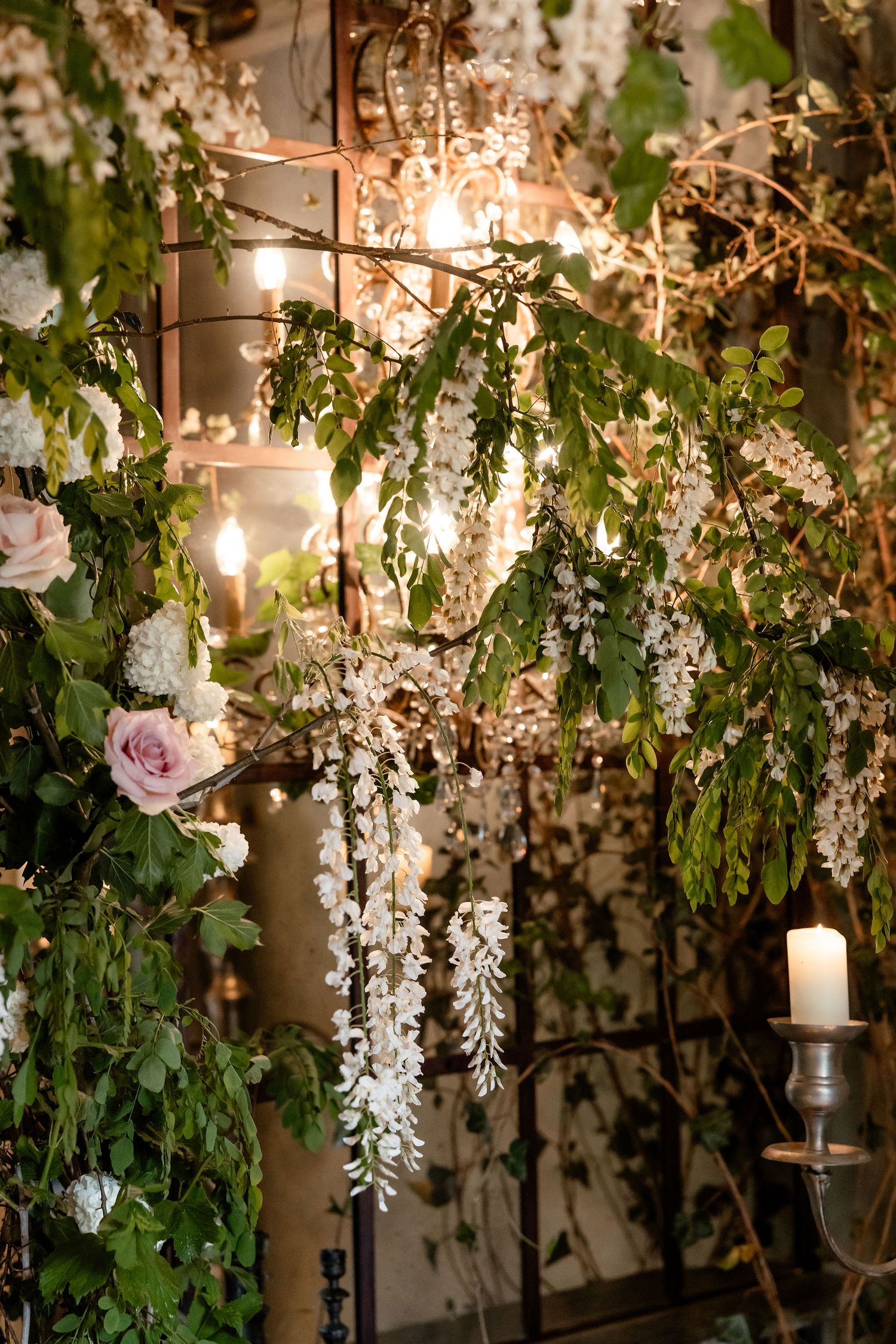 Romantic garden setting with hanging white wisteria flowers, greenery, pink roses and glowing candles creating ambient lighting.