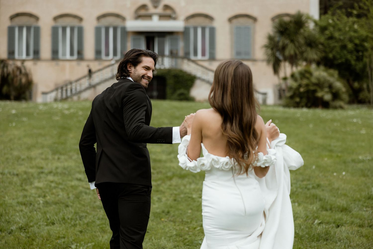 A couple celebrates their wedding day on a grassy lawn outside an elegant historic mansion.