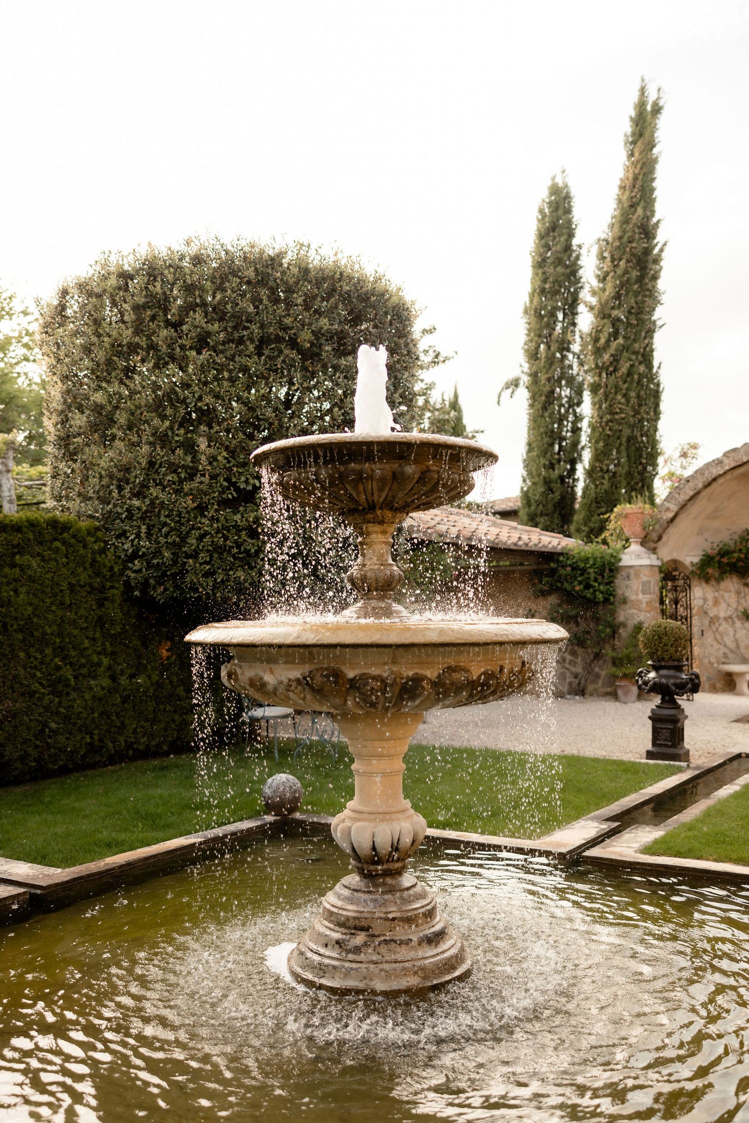 A classical tiered fountain sprays water in an elegant garden setting with tall cypress trees and manicured hedges.