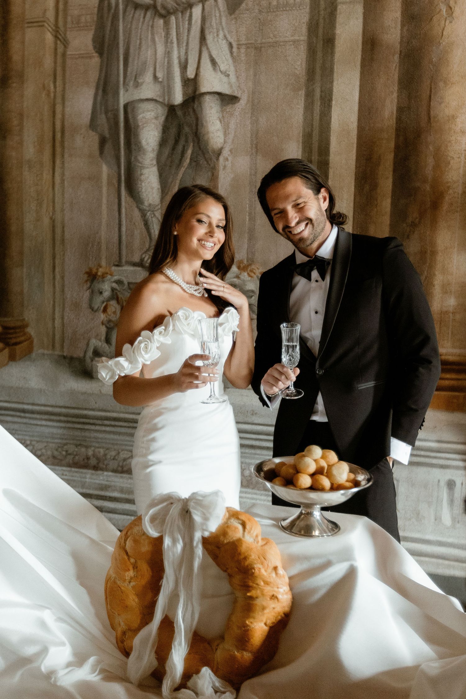 Elegant couple in formal wedding attire toasting with champagne glasses beside a decorative statue and pastry display.