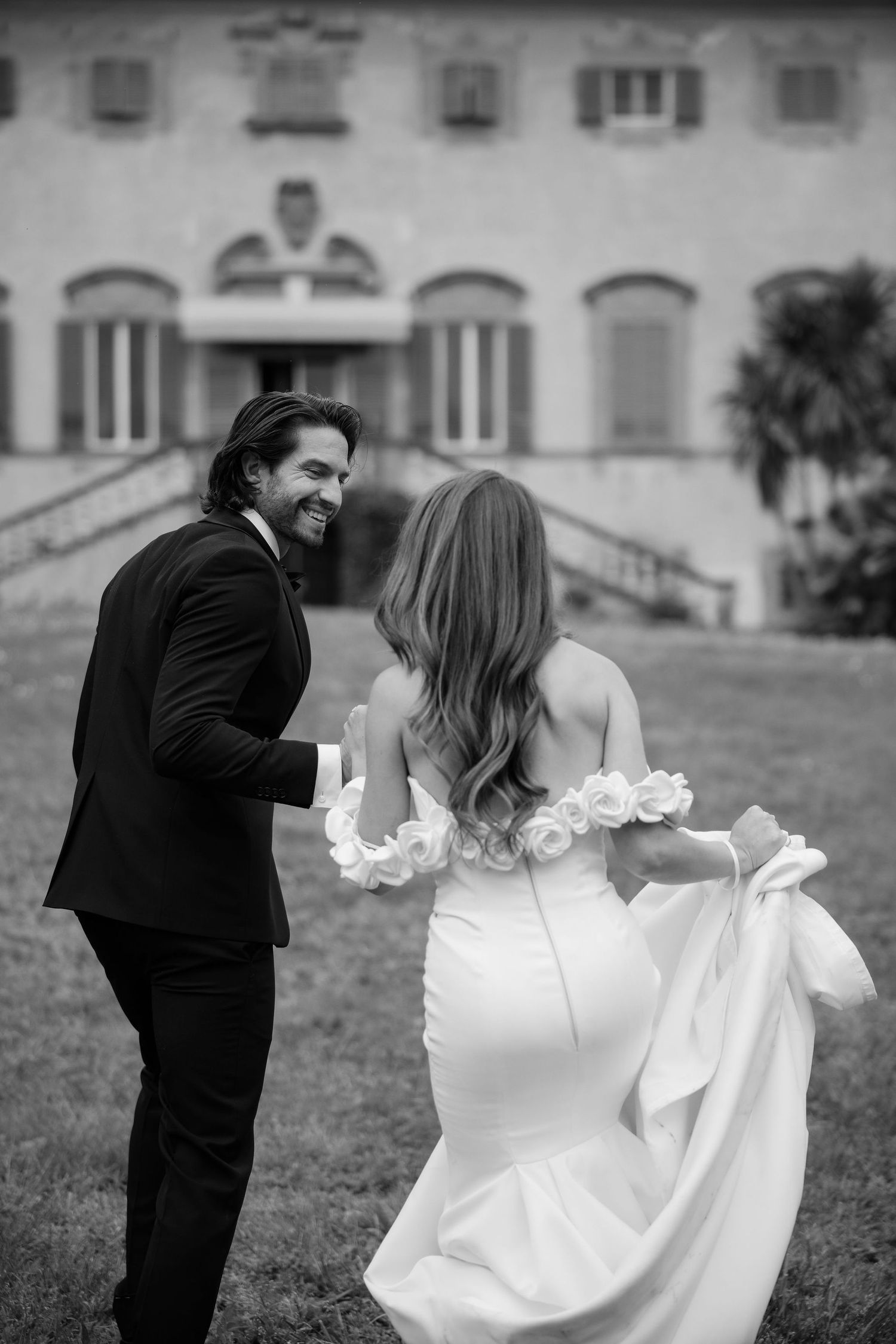 A couple shares a joyful moment in black and white as the bride holds her flowing white dress on the mansion grounds.