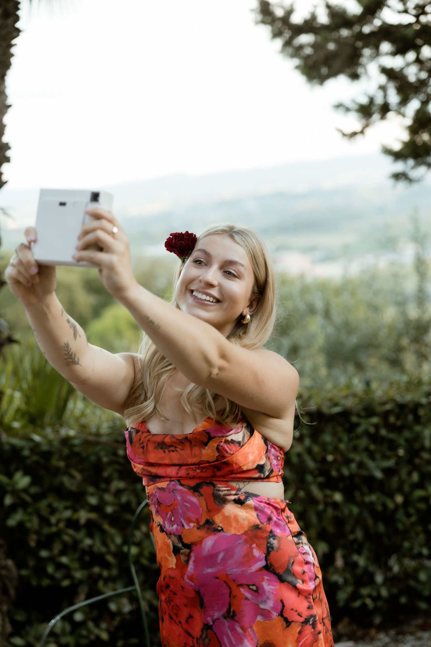 A person in a vibrant floral dress joyfully takes a selfie outdoors on a sunny day.