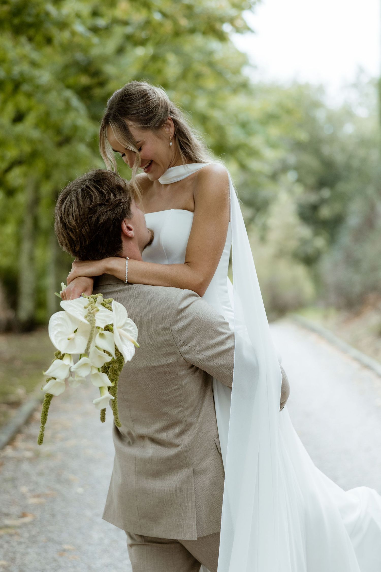 A romantic wedding couple embraces on a tree-lined path while holding white floral bouquet.