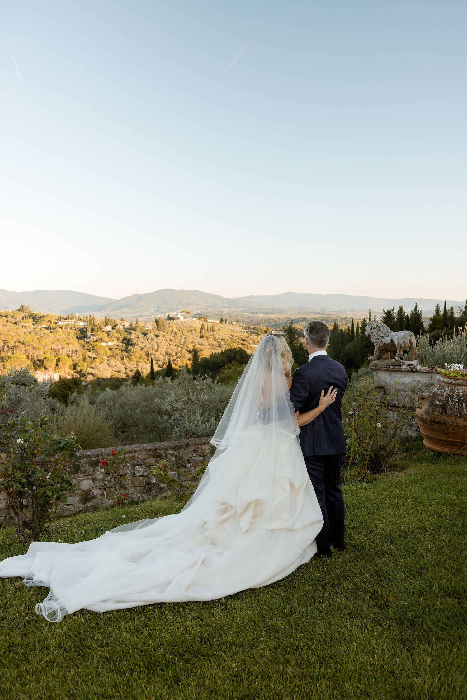 A couple in wedding attire stands on a grassy hill overlooking a scenic countryside landscape at sunset.