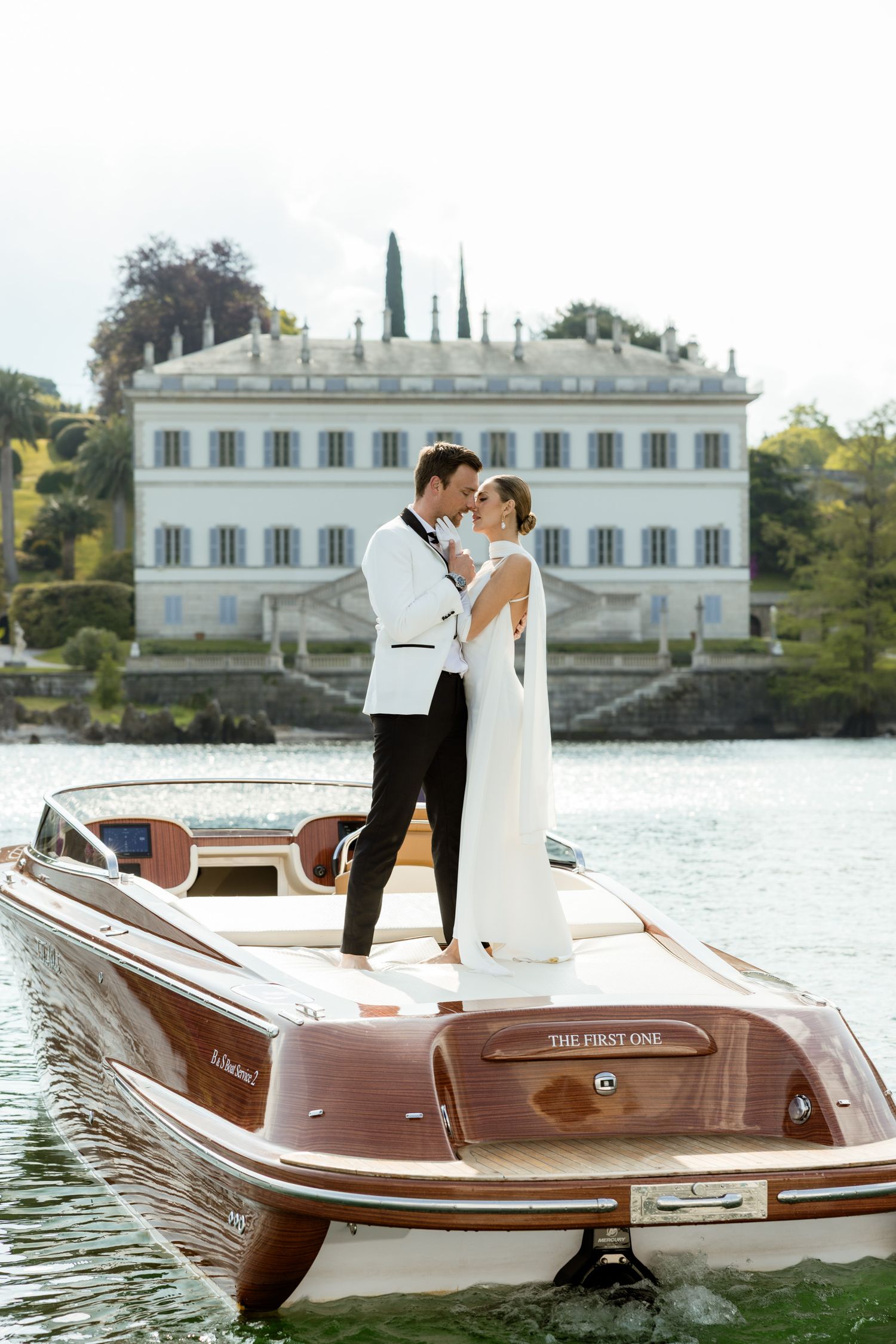 Bride and groom share romantic moment on wooden boat with elegant lakeside villa backdrop.