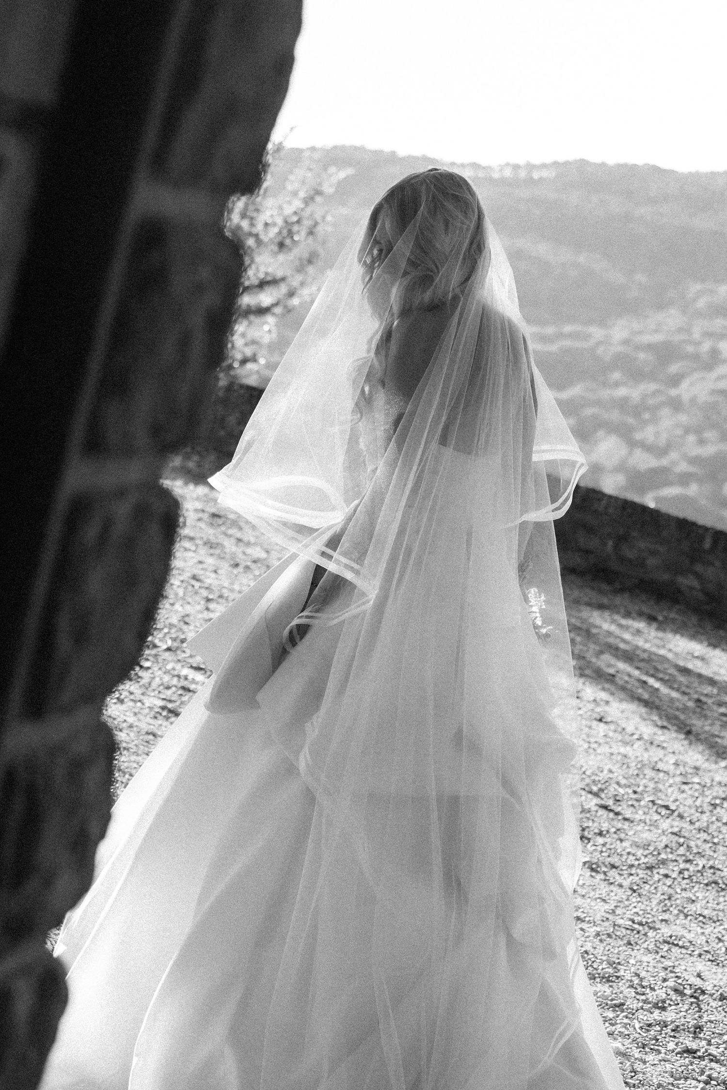 A bride in a flowing white wedding veil stands silhouetted against a scenic mountain landscape in black and white.